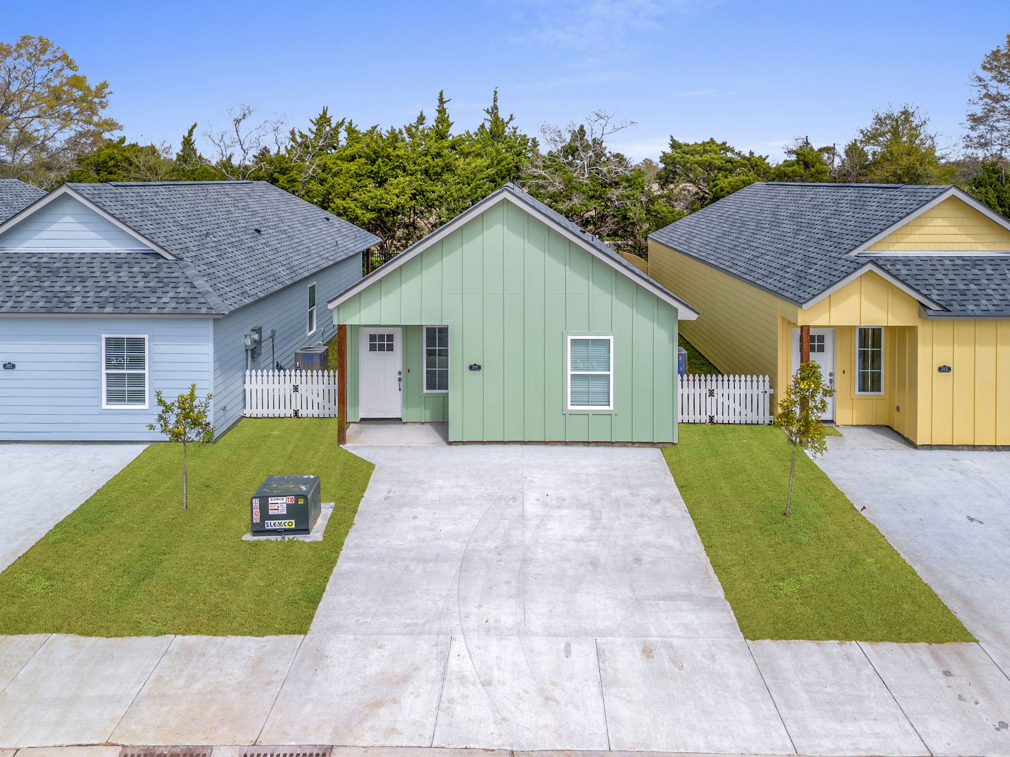 Aerial view of three single-story houses in a row: one is light blue, another is green, and the third is yellow. Each house features a well-maintained front yard with young trees and a concrete driveway. There's a utility box in the foreground, and the background includes green foliage and a clear blue sky.