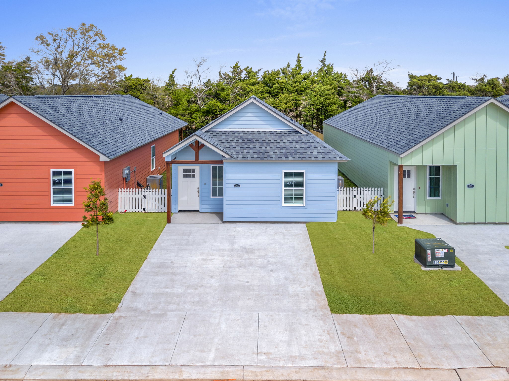 Three colorful single-story houses arranged in a row. The left house is red, the middle one is blue, and the right one is green. Each house features a porch, and a neatly manicured lawn is in front. A driveway connects the houses, and trees can be seen in the background under a clear blue sky.