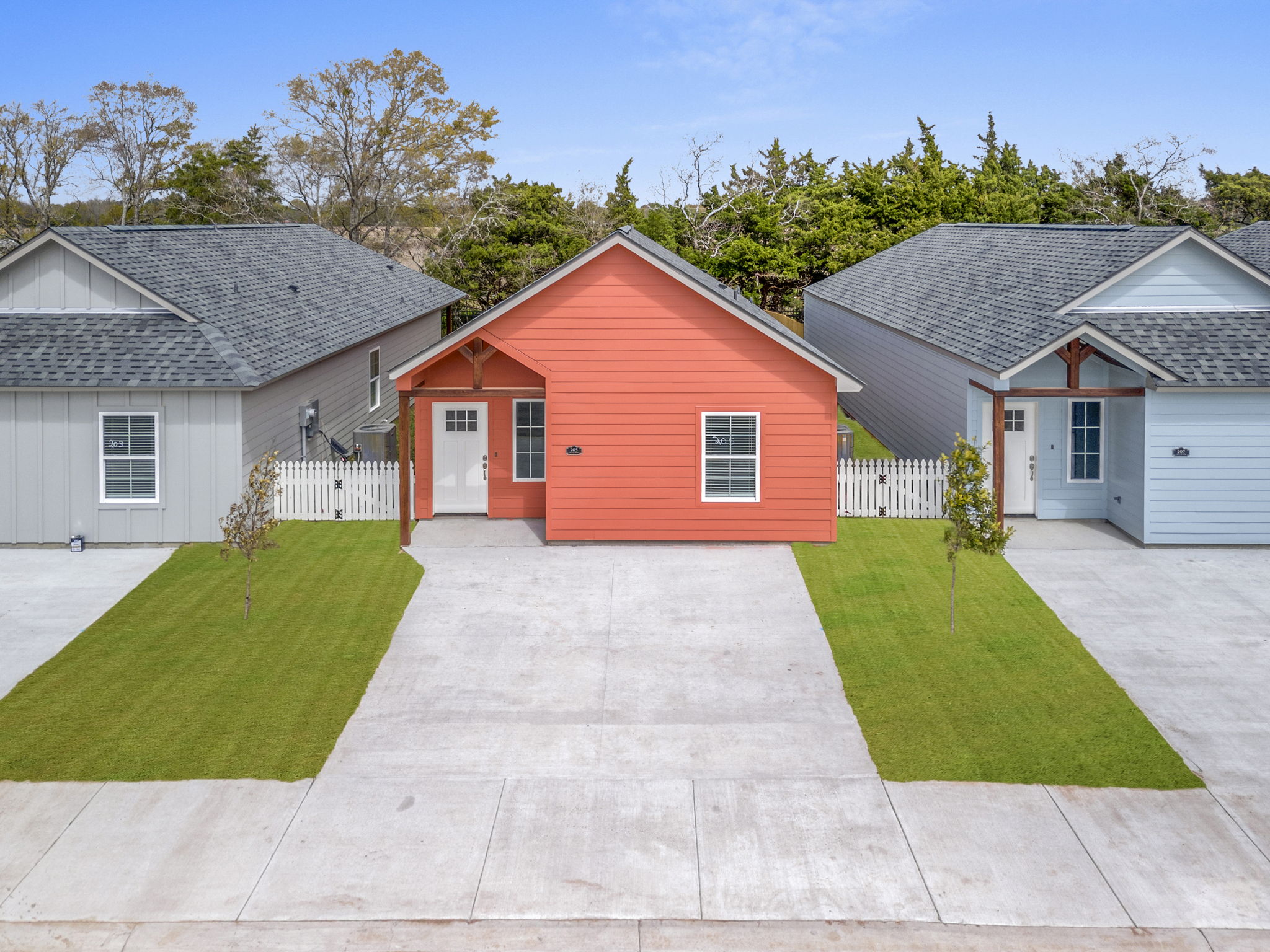 Aerial view of three modern single-family homes with a landscaped lawn. The center house is painted orange, while the two adjacent homes are gray and light blue. Each house features a front porch and is separated by driveways, surrounded by greenery in the background.