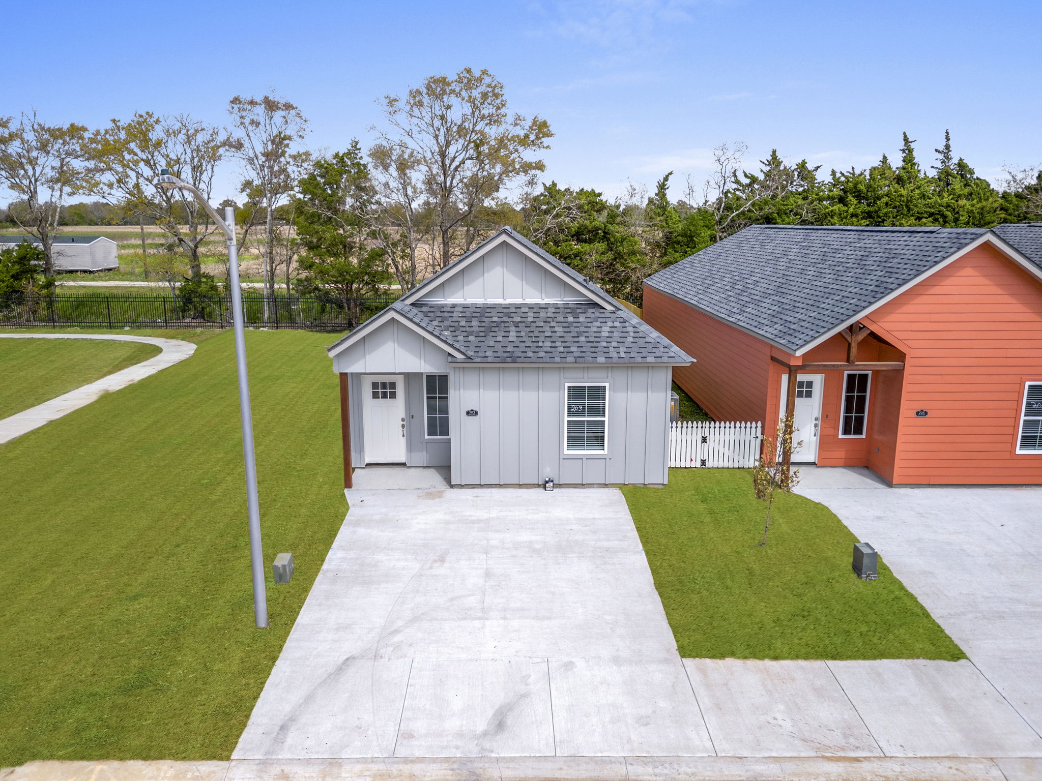 Aerial view of two single-story houses, one gray with a sloped roof and one orange, situated next to a neatly manicured lawn. A concrete driveway leads to the homes, with a light pole nearby. In the background, trees and open fields are visible under a clear blue sky.
