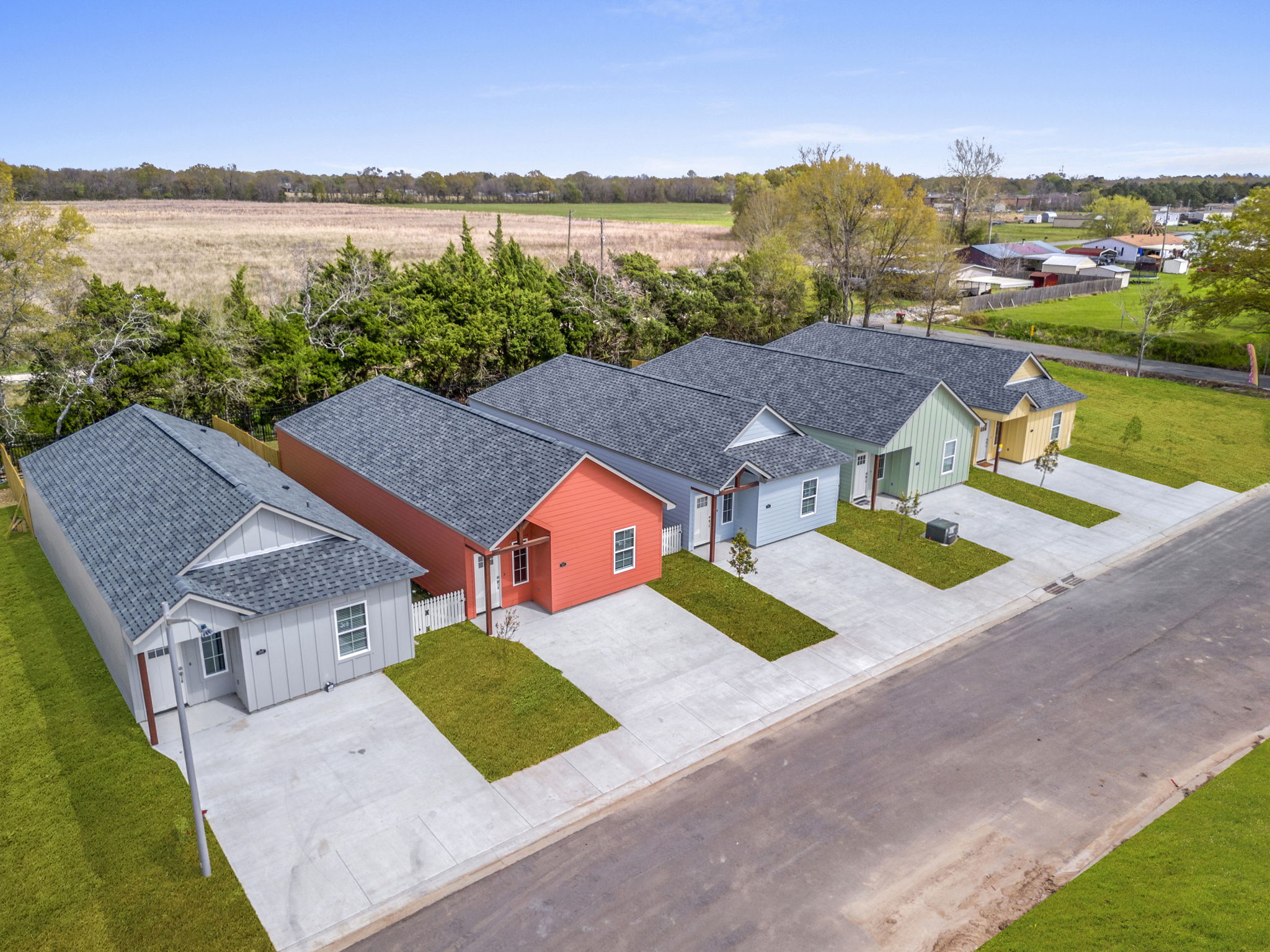 Aerial view of a row of colorful single-family homes with sloped roofs. Each house is a different color, including pink, green, and orange, situated along a paved street. Lush greenery and an open field are in the background, providing a tranquil suburban setting.