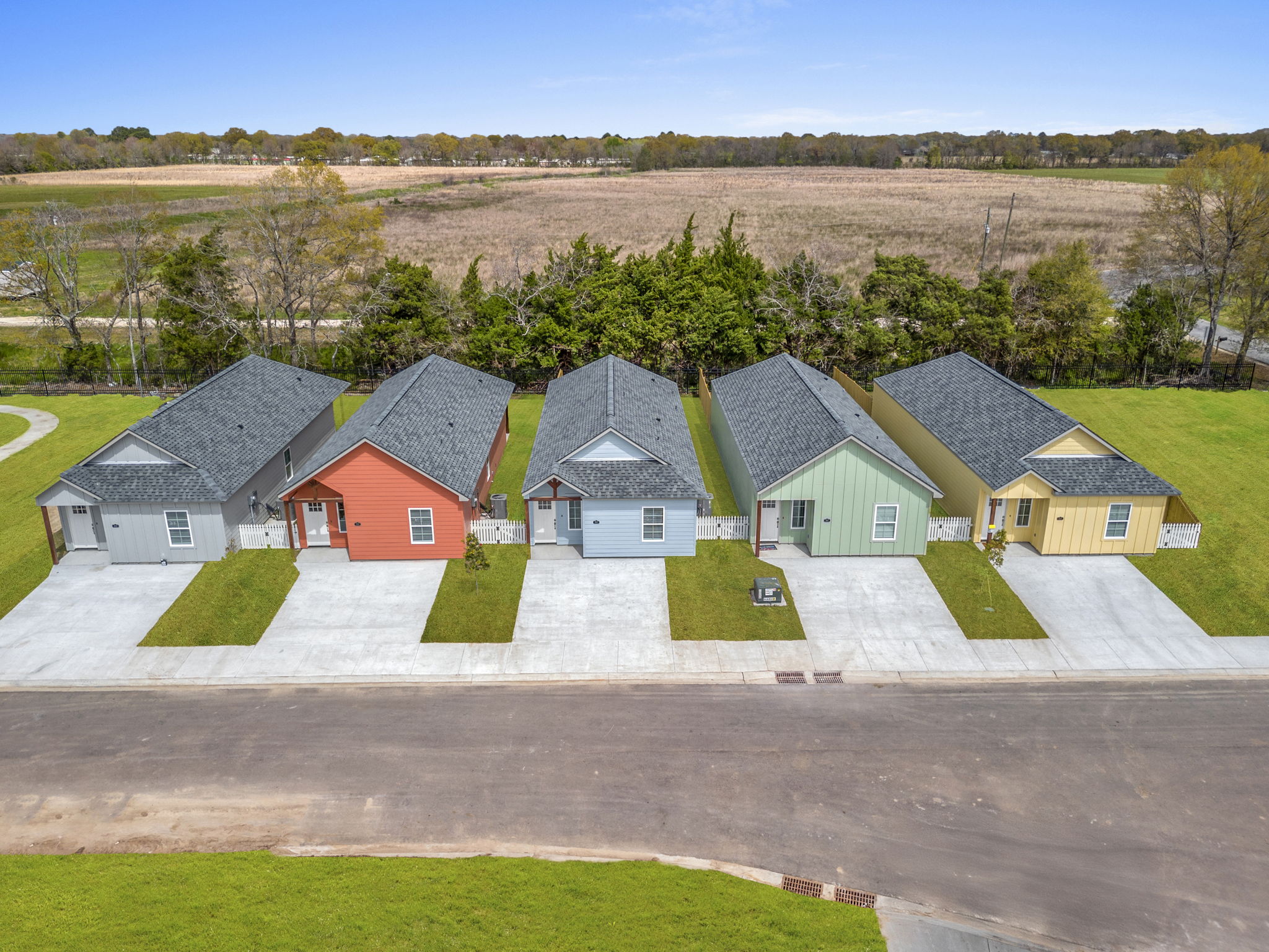 A row of colorful houses with gabled roofs sits along a newly paved road. The homes are arranged neatly, featuring varying shades of red, gray, green, and yellow. Lush green grass surrounds the houses, and a field is visible in the background under a clear blue sky.