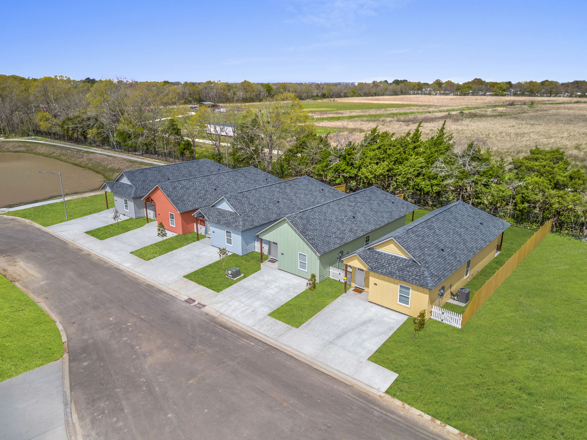 Aerial view of a row of colorful single-story houses on a quiet street. The houses are in shades of red, green, yellow, and gray, with manicured lawns and small trees. In the background, there are open fields and a clear blue sky, creating a serene suburban atmosphere.