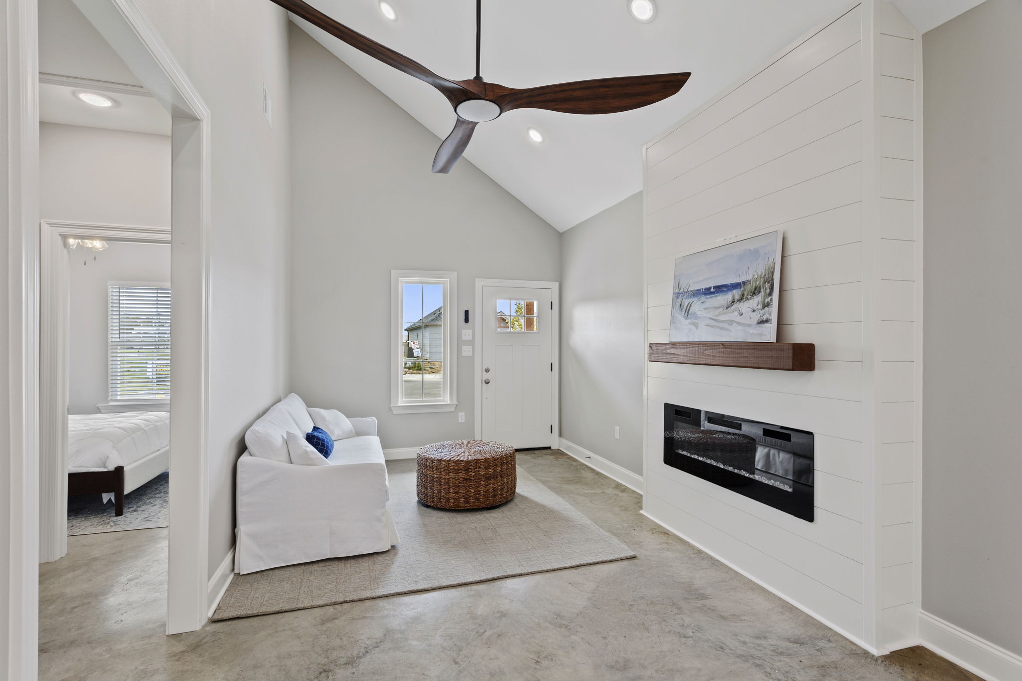 A modern living room with a high ceiling, featuring a white sofa, a round wicker coffee table, and a sleek fireplace. Natural light floods in through large windows, and there’s a decorative painting on the wall. The floor has a light concrete finish, creating a spacious and airy atmosphere.
