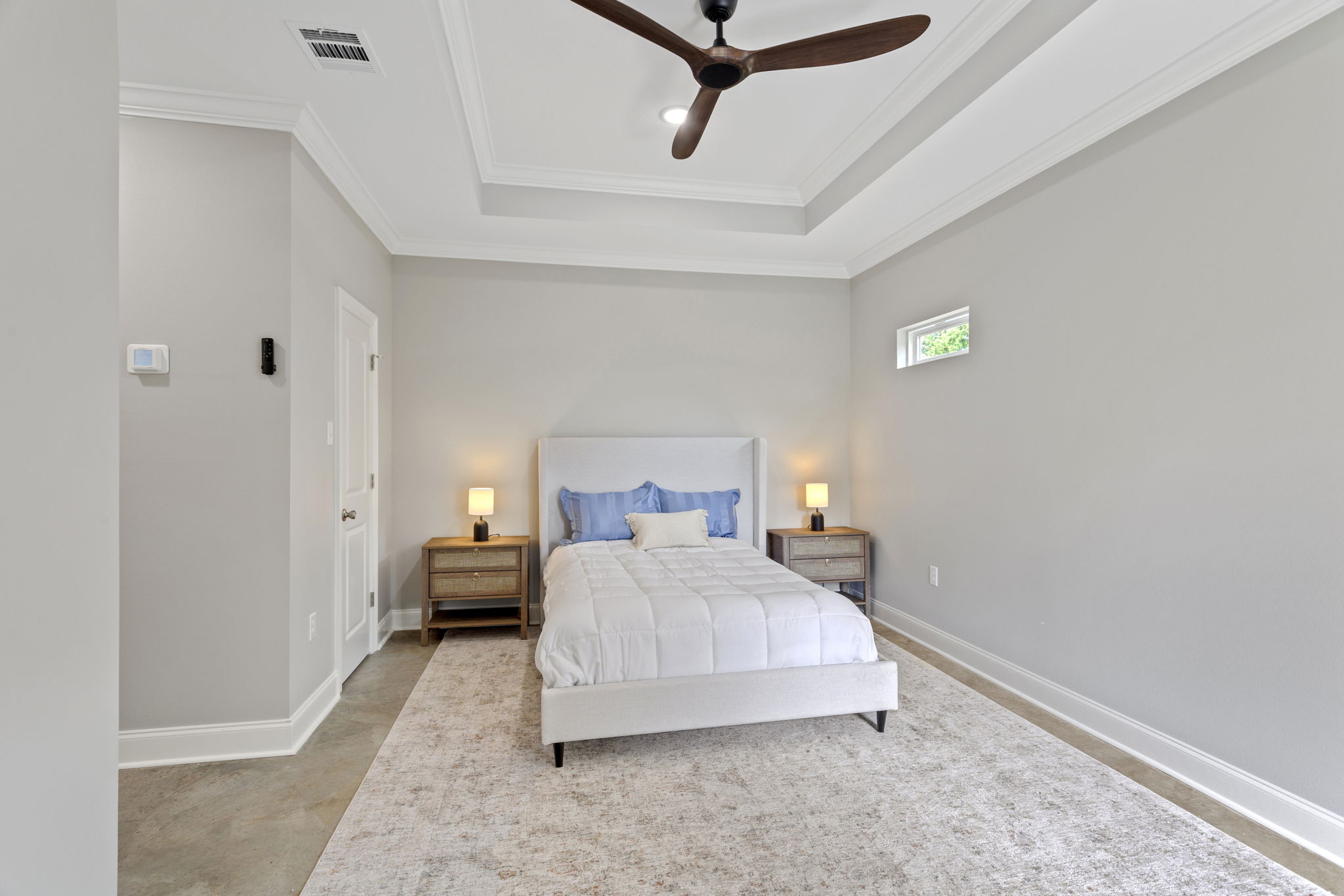A modern bedroom featuring a white bed with a light blue accent pillow, two bedside tables with lamps, and a large area rug. The ceiling has a decorative design and a ceiling fan. The walls are painted light gray, creating a calm and inviting atmosphere. A small window allows natural light to enter.