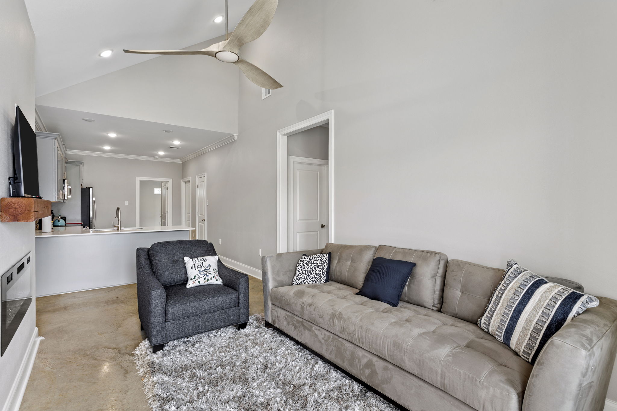 A bright living room featuring a light gray sofa with decorative pillows and a black armchair. The space has a ceiling fan and is adorned with a plush, light-colored rug. In the background, a modern kitchen area is visible, with sleek cabinetry and appliances. The walls are painted a soft neutral tone.