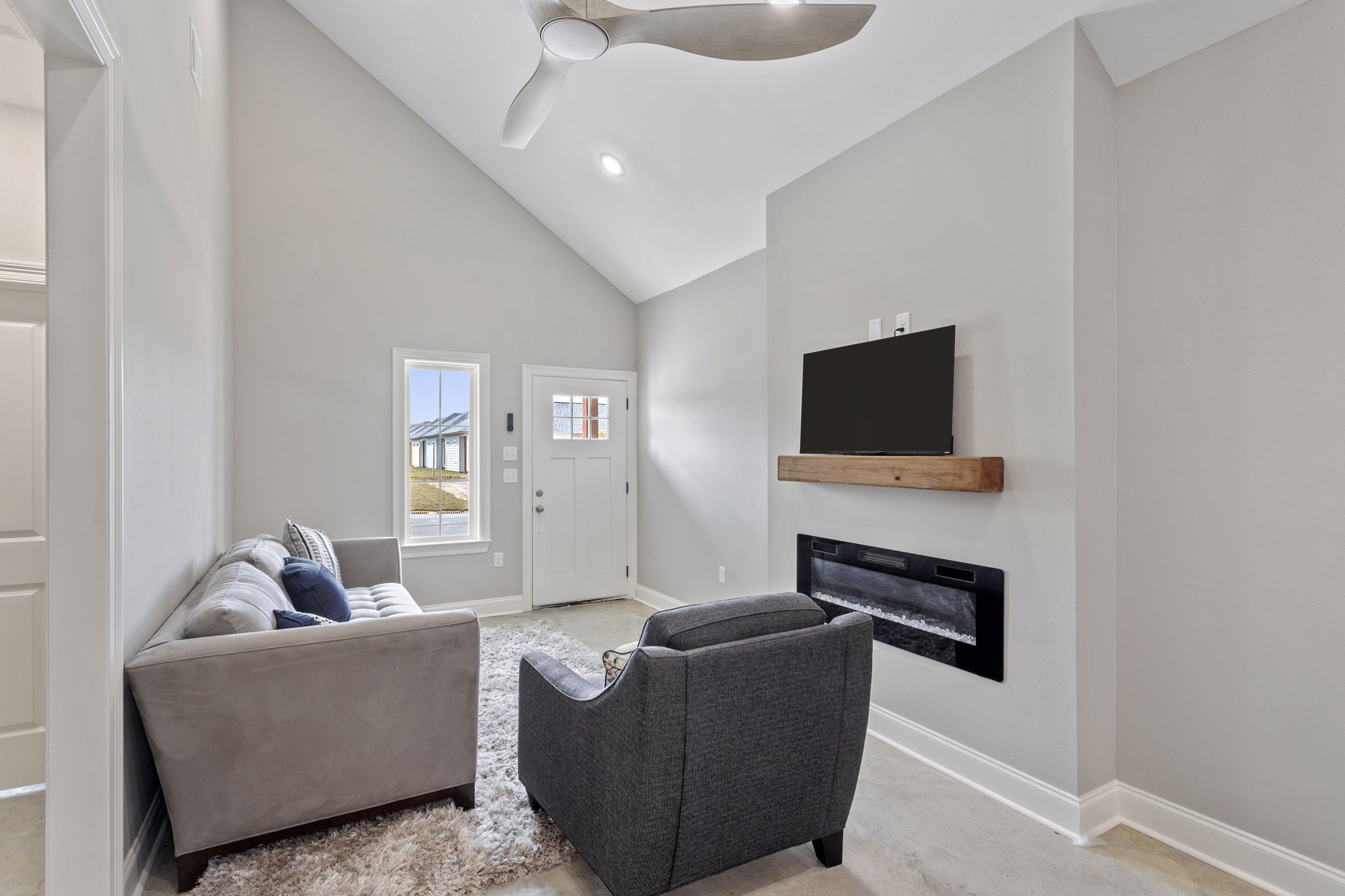Modern living room with a gray sofa and a dark gray armchair. Features include a sleek ceiling fan, a mounted flat-screen TV, and a minimalist electric fireplace. Large windows provide natural light, and the walls are painted in a soft neutral tone, creating a cozy atmosphere.