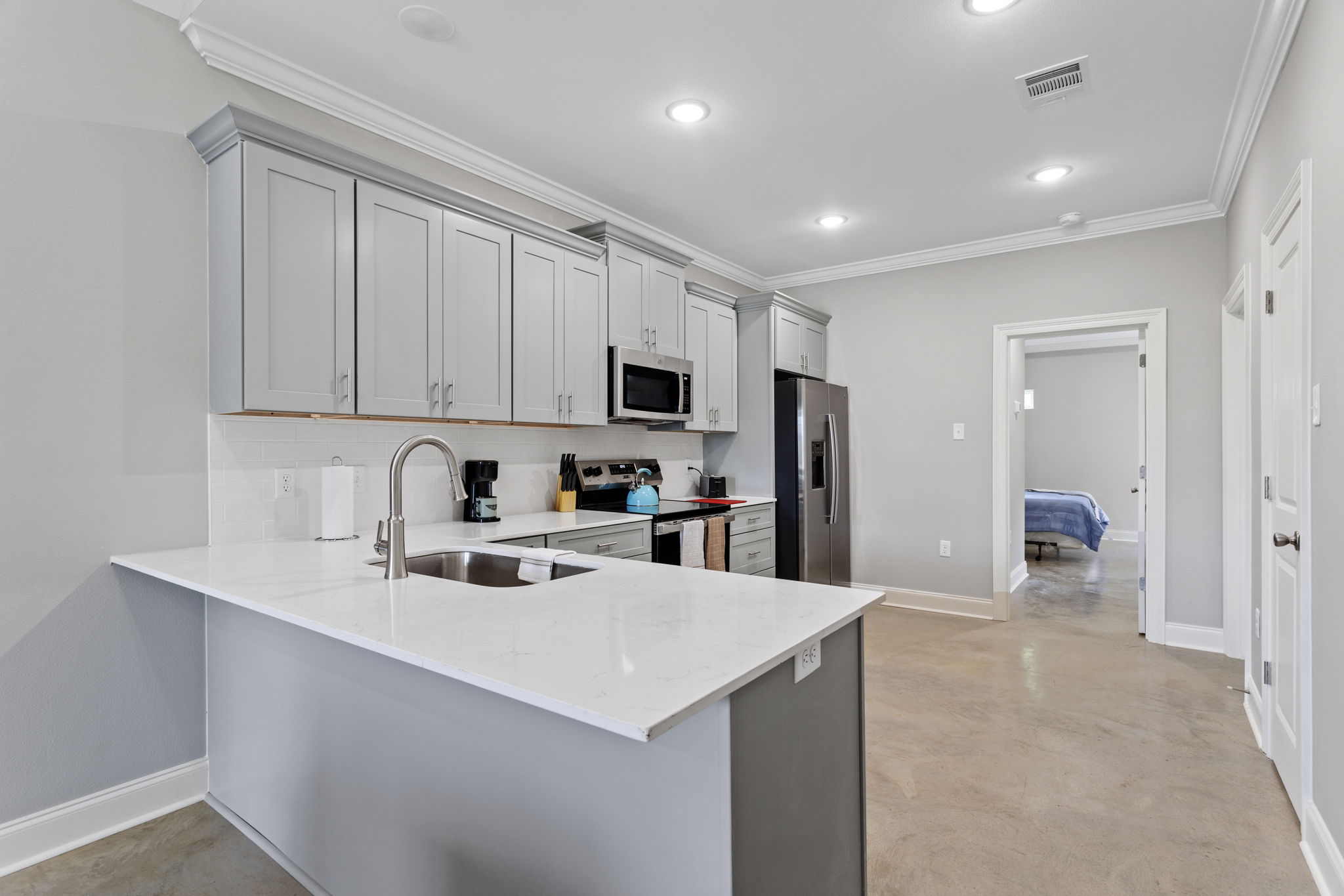 Modern kitchen featuring gray cabinetry, stainless steel appliances, and a large island with a sink. Bright lighting illuminates the space, highlighting a clean and contemporary design. In the background, a doorway leads to a bedroom area, visible through an open door.