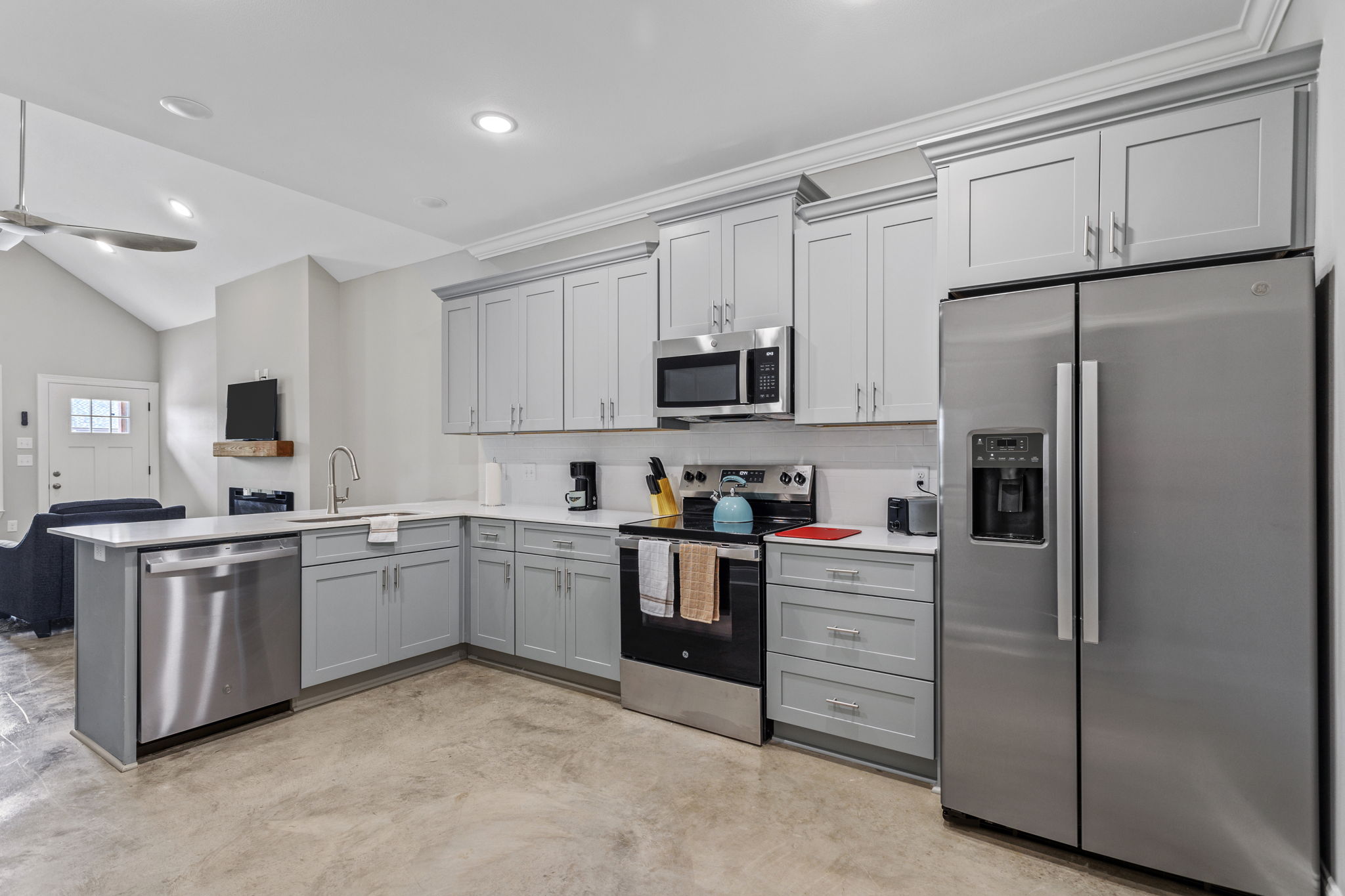 Modern kitchen featuring light gray cabinets, stainless steel appliances, and a spacious layout. The kitchen includes a refrigerator, microwave, and dishwasher, with a countertop area for food preparation. Bright lighting enhances the open atmosphere, making it a welcoming space for cooking and dining.