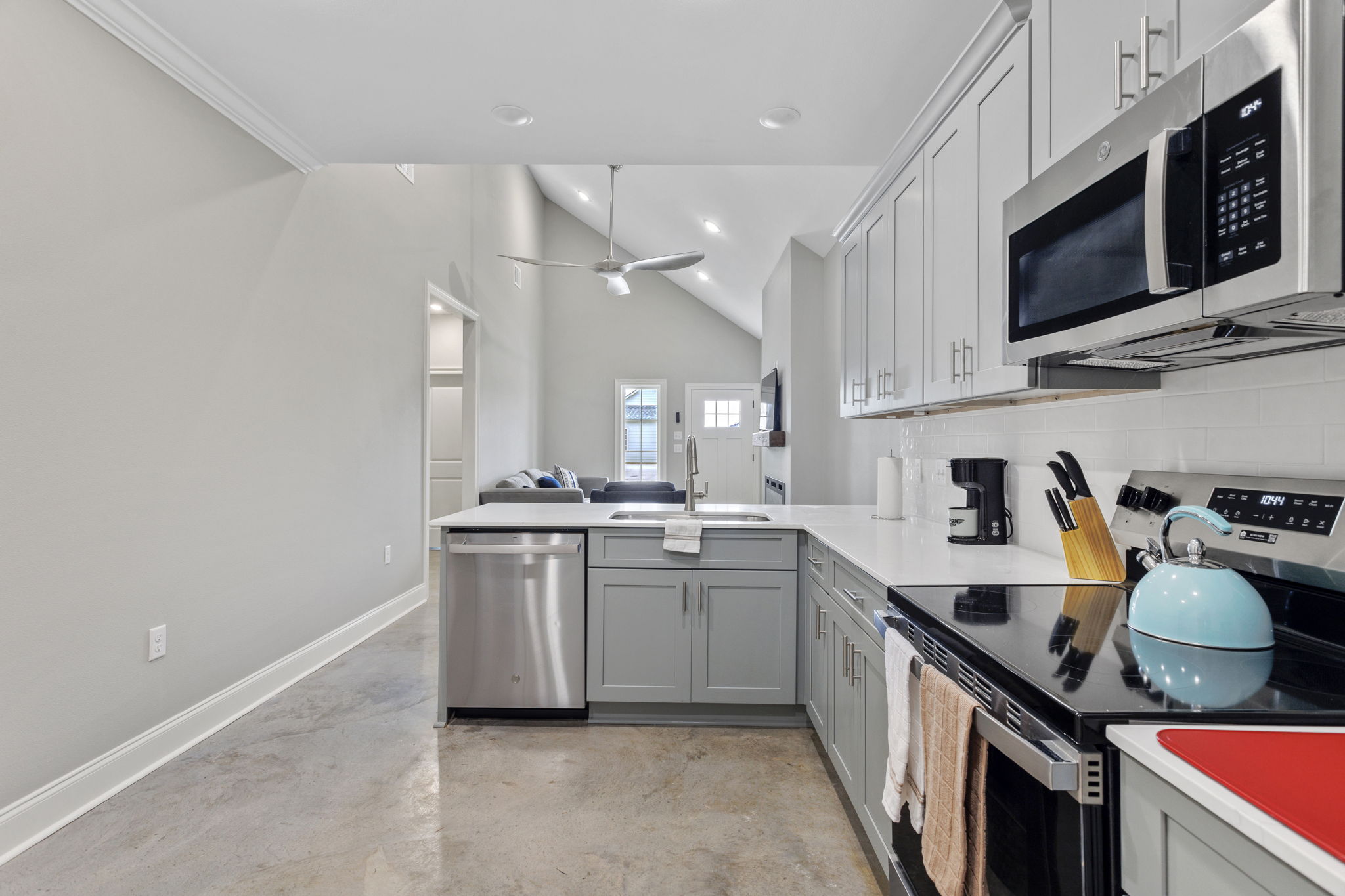 Modern kitchen featuring gray cabinets, stainless steel appliances, and a turquoise kettle. The space includes a dishwasher and an oven, with a minimalist design and natural light coming from an adjoining living area. The floor is polished concrete, and there is a ceiling fan visible.
