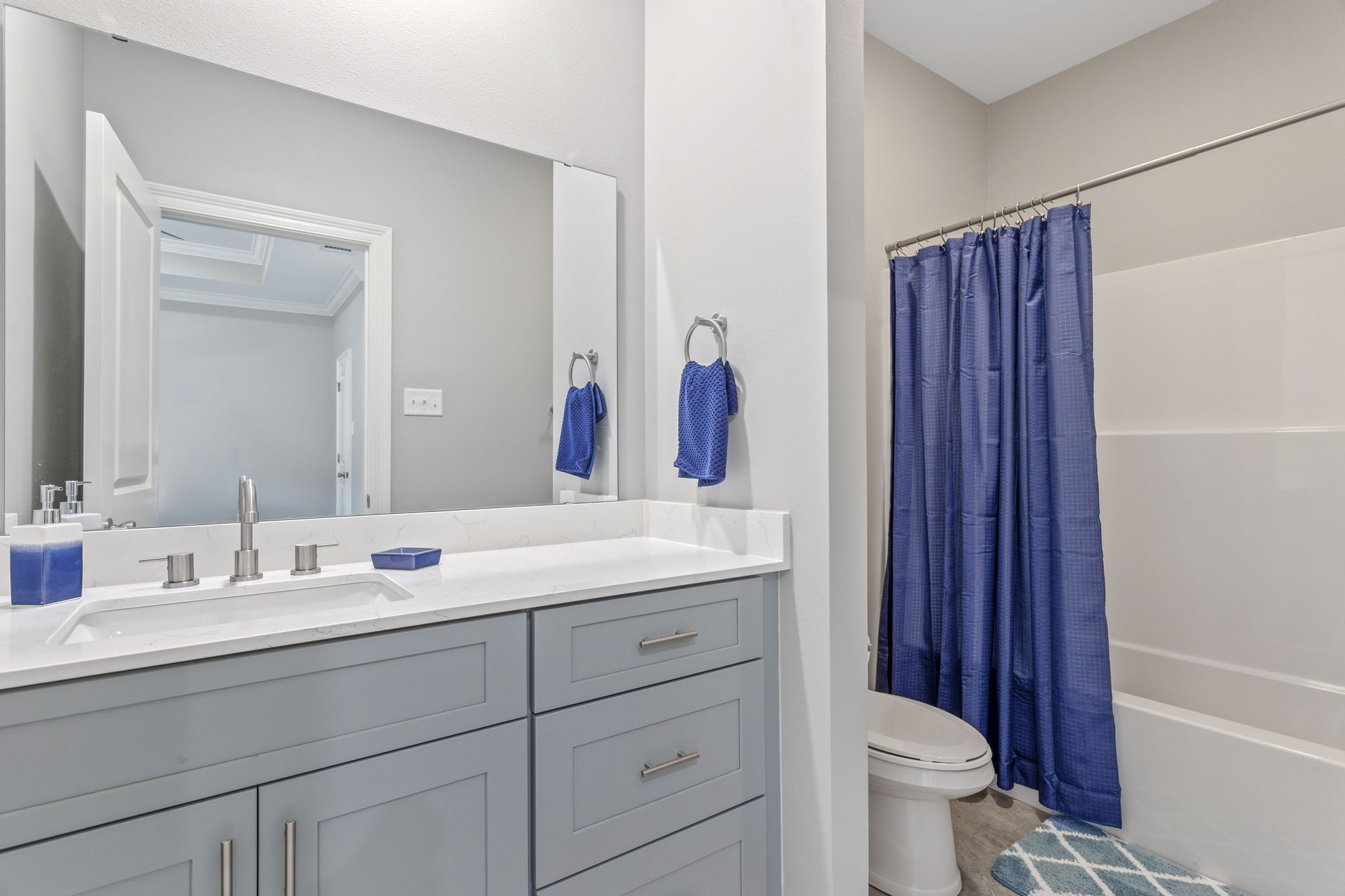 A modern bathroom featuring a light gray vanity with a white countertop, a large mirror, and two blue decorative towels hanging on towel rings. There is a blue shower curtain covering the bathtub, and a beige rug on the floor. The walls are painted in a soft gray tone.