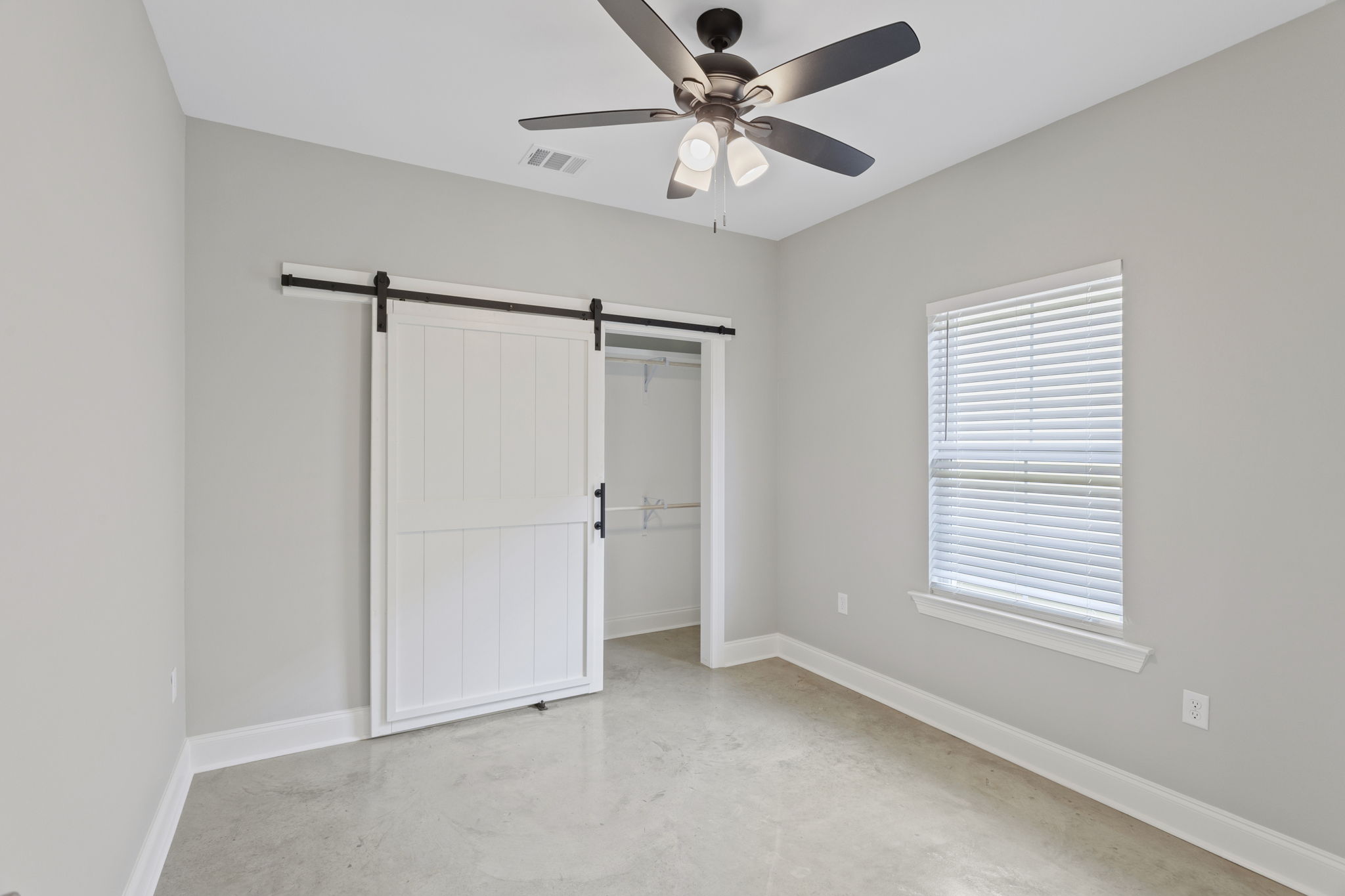 A spacious, empty room with light gray walls, a ceiling fan with five blades, and polished concrete flooring. There is a white sliding barn door leading to a closet, and a window with white blinds allowing natural light into the space.