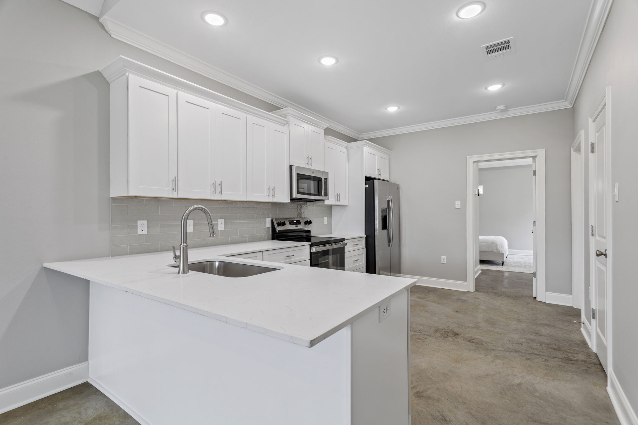 Modern kitchen featuring white cabinetry, stainless steel appliances including a microwave and refrigerator, an island with a sink, and polished concrete flooring. The space is bright with recessed lighting and has an open layout leading to another room.