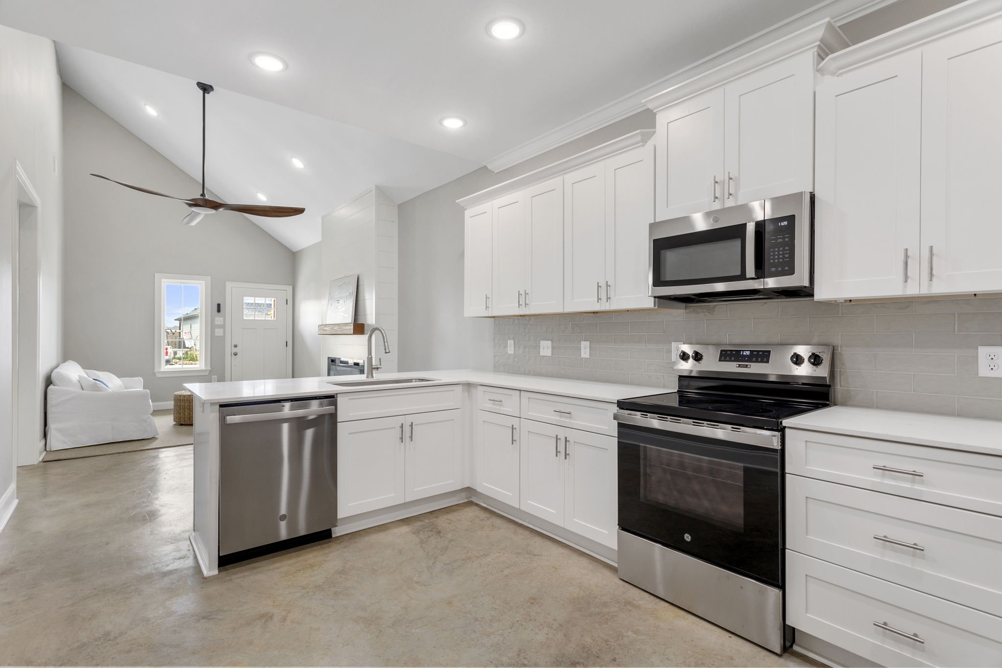 Modern kitchen with white cabinetry, stainless steel appliances, and a large island. The space features a clean design with concrete flooring and recessed lighting. A comfortable living area is visible in the background, showcasing a bright and airy atmosphere. Natural light streams in through nearby windows.