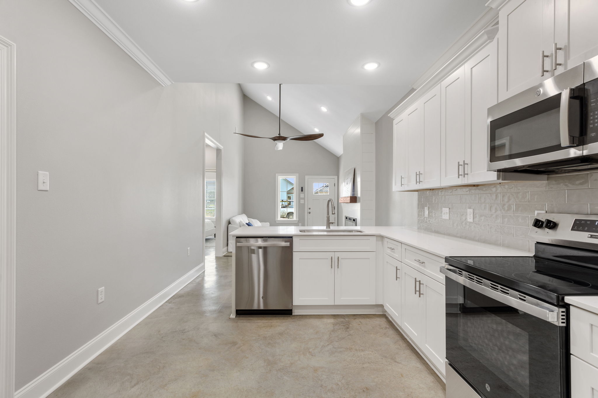 A modern kitchen featuring white cabinetry, stainless steel appliances, and a spacious countertop. The open layout includes a dining area visible in the background, along with a ceiling fan and recessed lighting, creating a bright and airy atmosphere. The floor has a polished concrete finish.