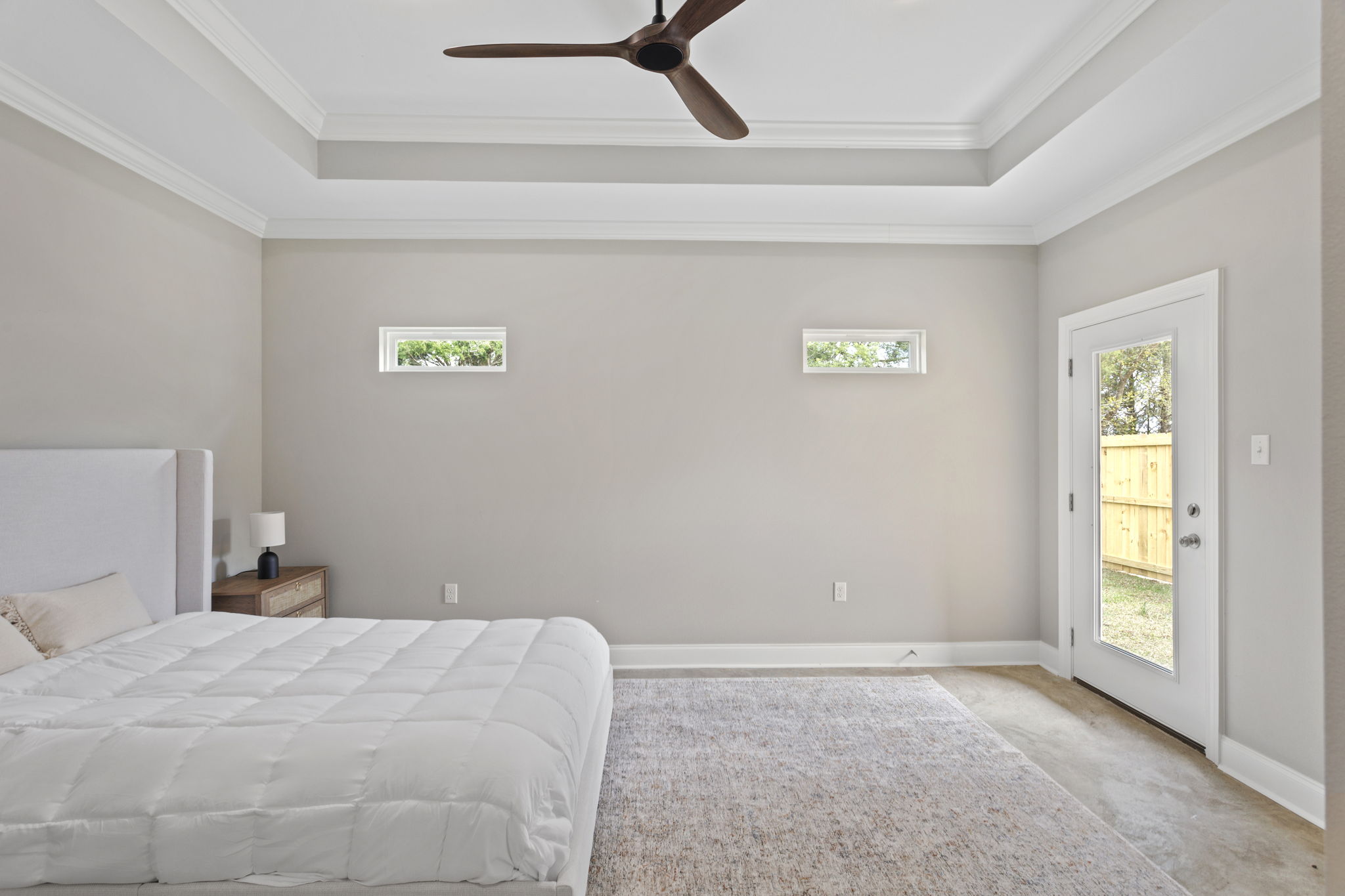A spacious, minimalist bedroom featuring a white bed with a quilt, a wooden nightstand with a lamp, and light-colored walls. Two small windows let in natural light, and a door leads to an outdoor area, enhancing the bright and airy atmosphere. A neutral-toned area rug covers the floor.