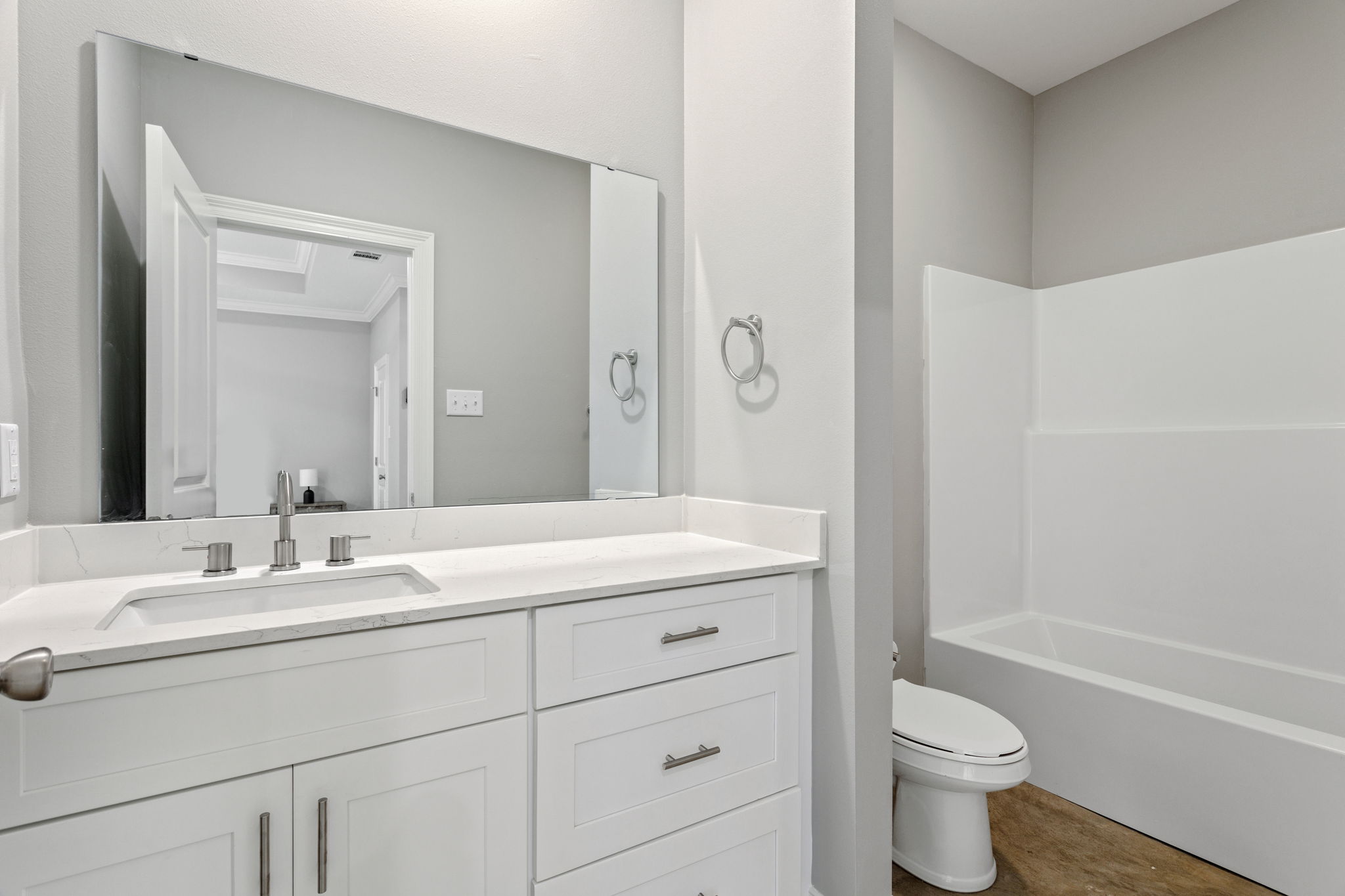 A modern bathroom featuring a white vanity with a sink and mirror, a towel ring, and a separate toilet area. The decor is minimalistic with light gray walls. Adjacent is a bathtub enclosed in a white surround. The space is well-lit and clean, reflecting a contemporary design.