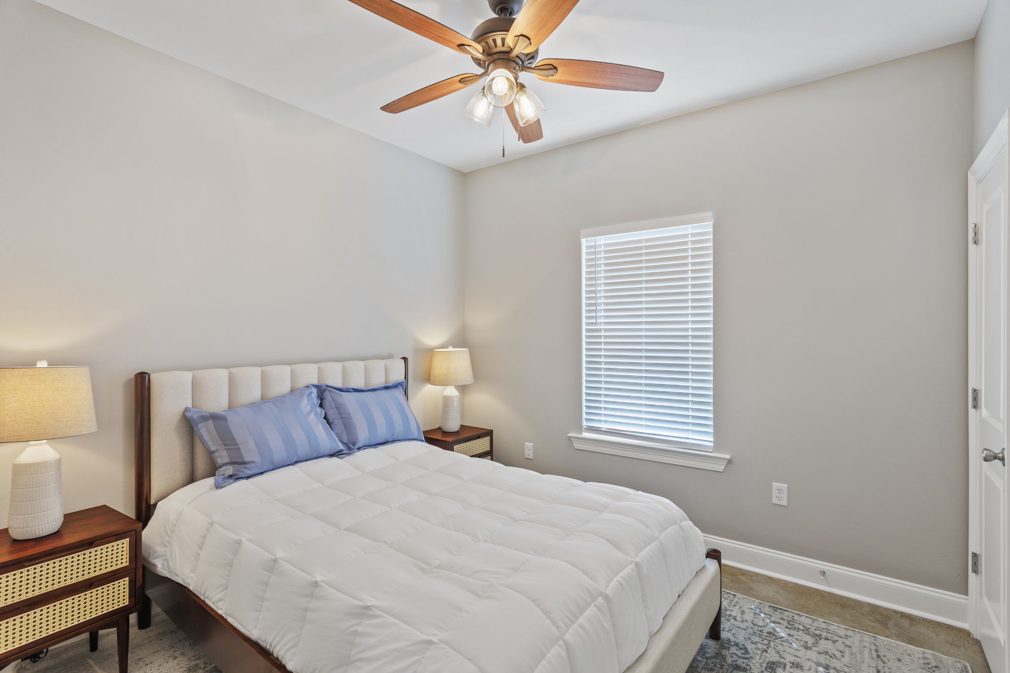 A cozy bedroom featuring a large bed with white bedding, two bedside lamps on wooden tables, and a ceiling fan. The room has light-colored walls and a window with blinds, allowing natural light to enter, creating a bright and inviting atmosphere.