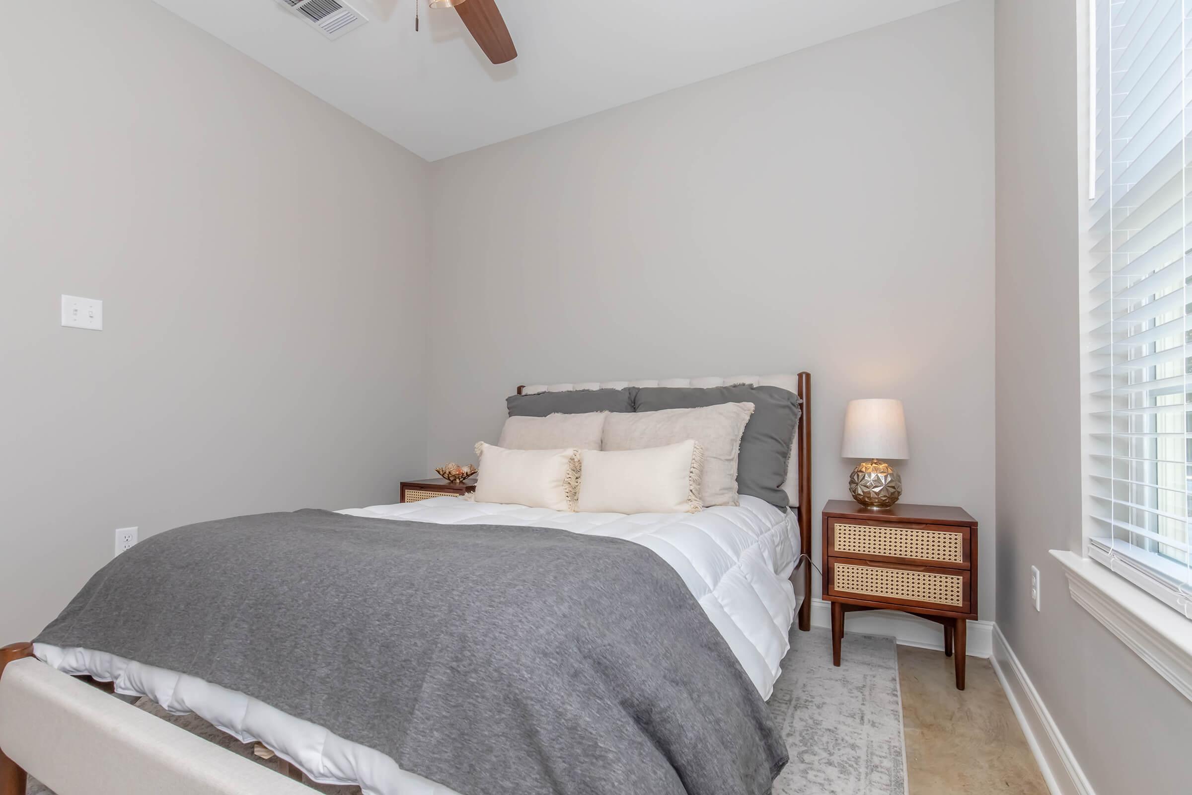 A cozy bedroom featuring a neatly made bed with gray bedding and decorative pillows. There is a small nightstand with a lamp beside the bed, and the walls are painted in a soft gray color. Natural light comes through a window, contributing to the warm and inviting atmosphere.