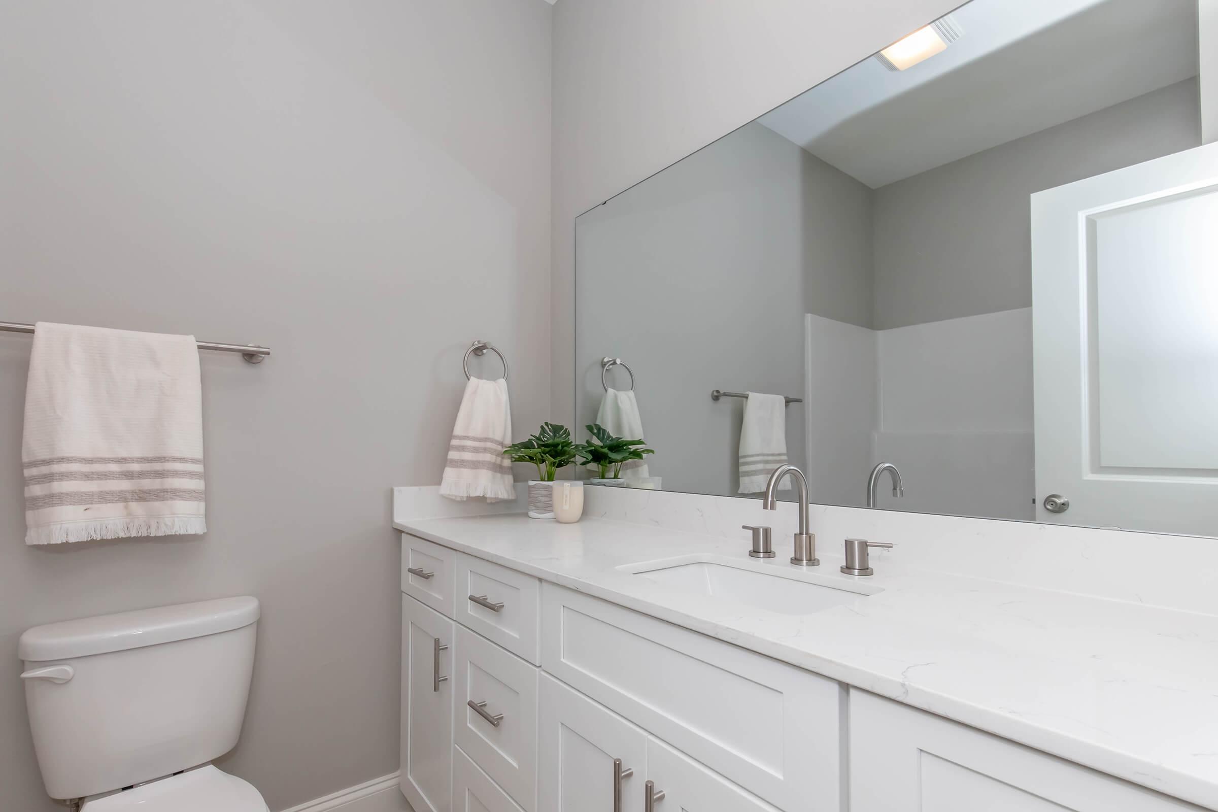 A modern bathroom featuring a white countertop with two sinks, a large mirror above, a light gray wall, a towel rack with two towels, and a small potted plant. A white toilet is visible on the left side. Bright and clean design emphasizes simplicity and elegance.