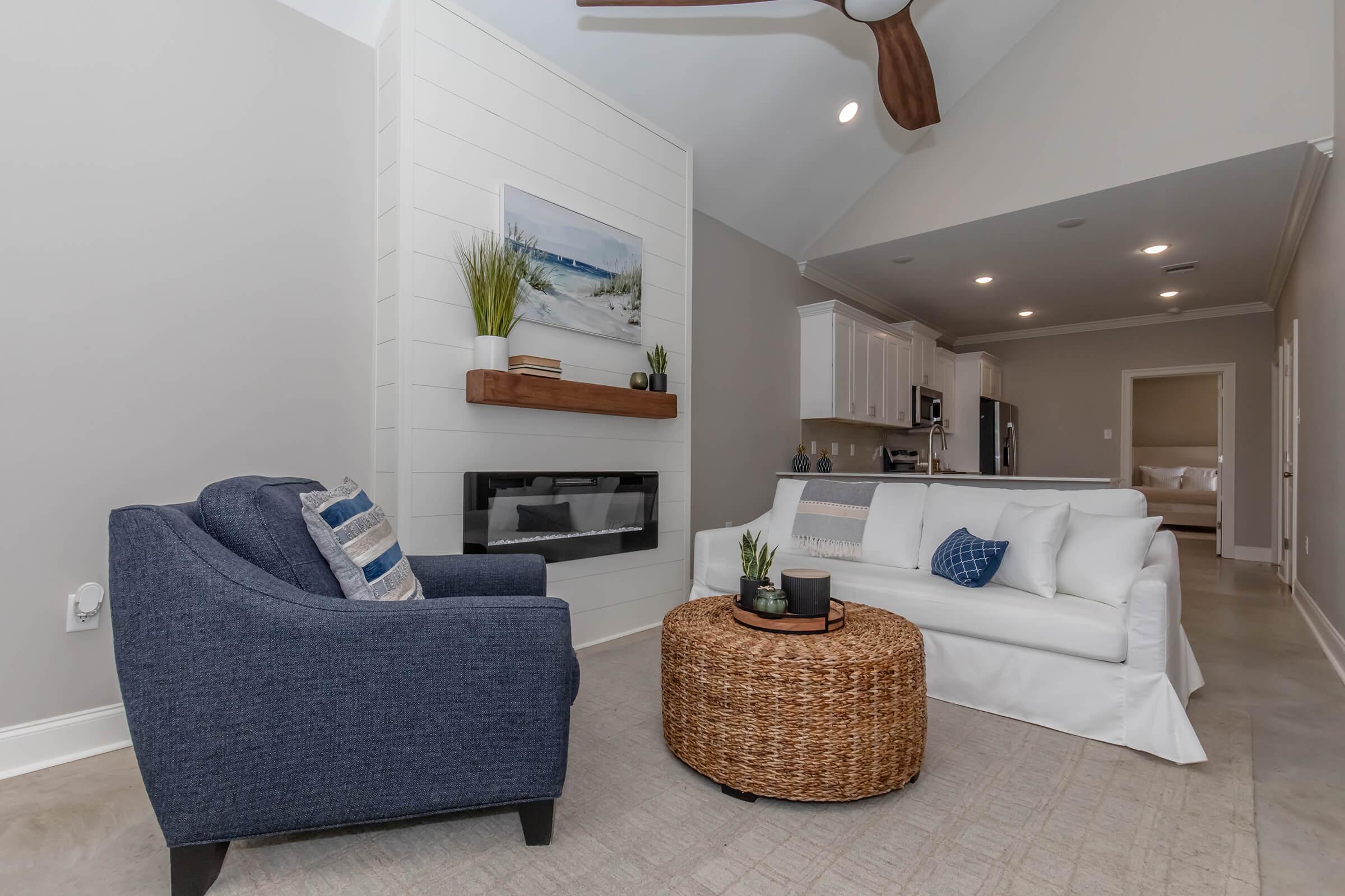 A cozy living room featuring a blue upholstered armchair, a white sofa with decorative pillows, and a round wicker coffee table. A modern wall-mounted fireplace is visible, along with a white shiplap accent wall. The room has neutral tones and a light, airy atmosphere, complemented by a kitchen area in the background.