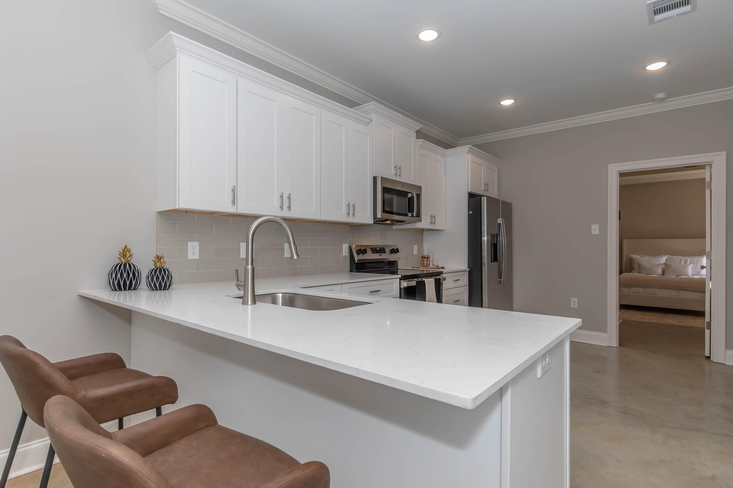 Modern kitchen featuring white cabinetry, stainless steel appliances, and a large island with a sink. Brown bar stools are positioned at the island. Soft lighting enhances the airy atmosphere. A doorway in the background leads to a living area with neutral-colored furnishings.