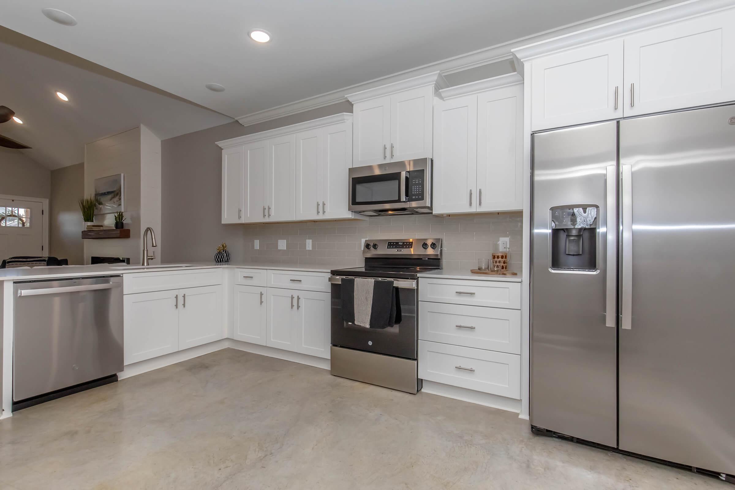 A modern kitchen featuring white cabinetry, stainless steel appliances including a microwave, oven, and refrigerator, and a large sink. The countertops are subtle and the flooring is polished concrete. Natural light fills the space, creating a bright and inviting atmosphere.