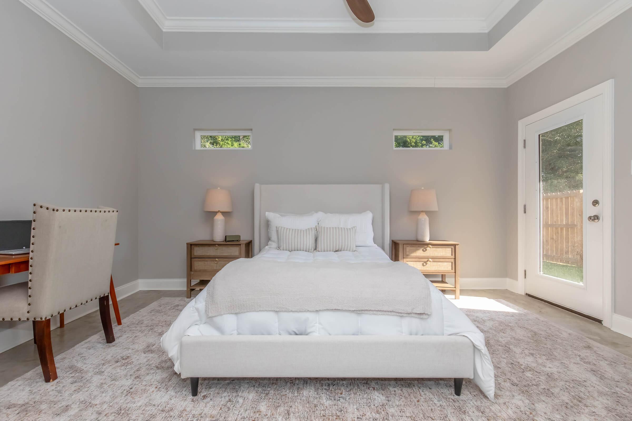 A modern bedroom featuring a white bed with a soft beige throw blanket, two bedside tables with lamps, and a cozy rug. A small desk is placed to the left, and there are large windows allowing natural light. The room has a neutral color palette, enhancing its serene atmosphere.