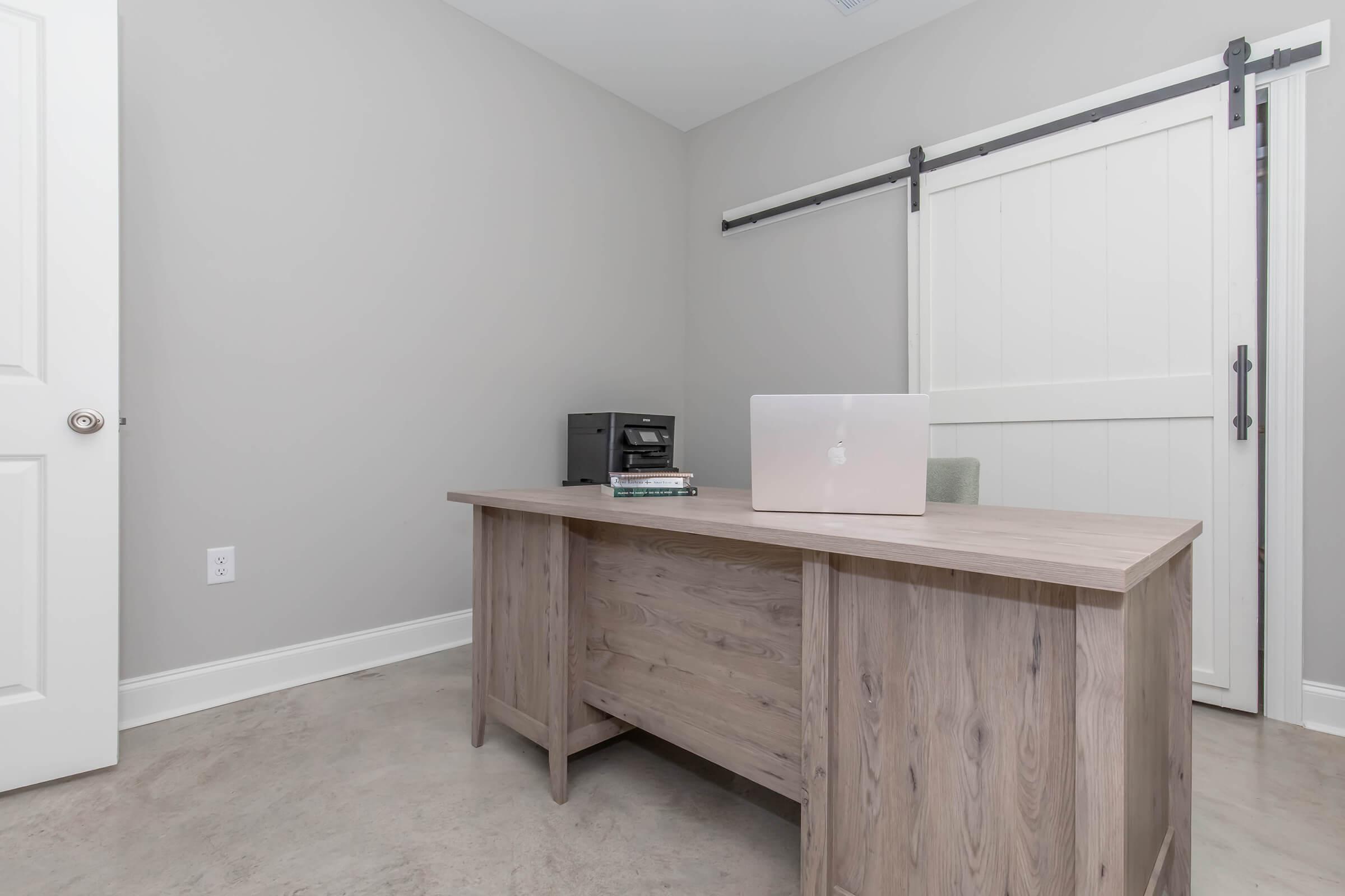 A modern office setup featuring a light wooden desk with a laptop on it, a stack of books, and a black printer. The room has gray walls and a white sliding barn door. Natural light enhances the minimalist design, creating a clean and inviting workspace atmosphere.
