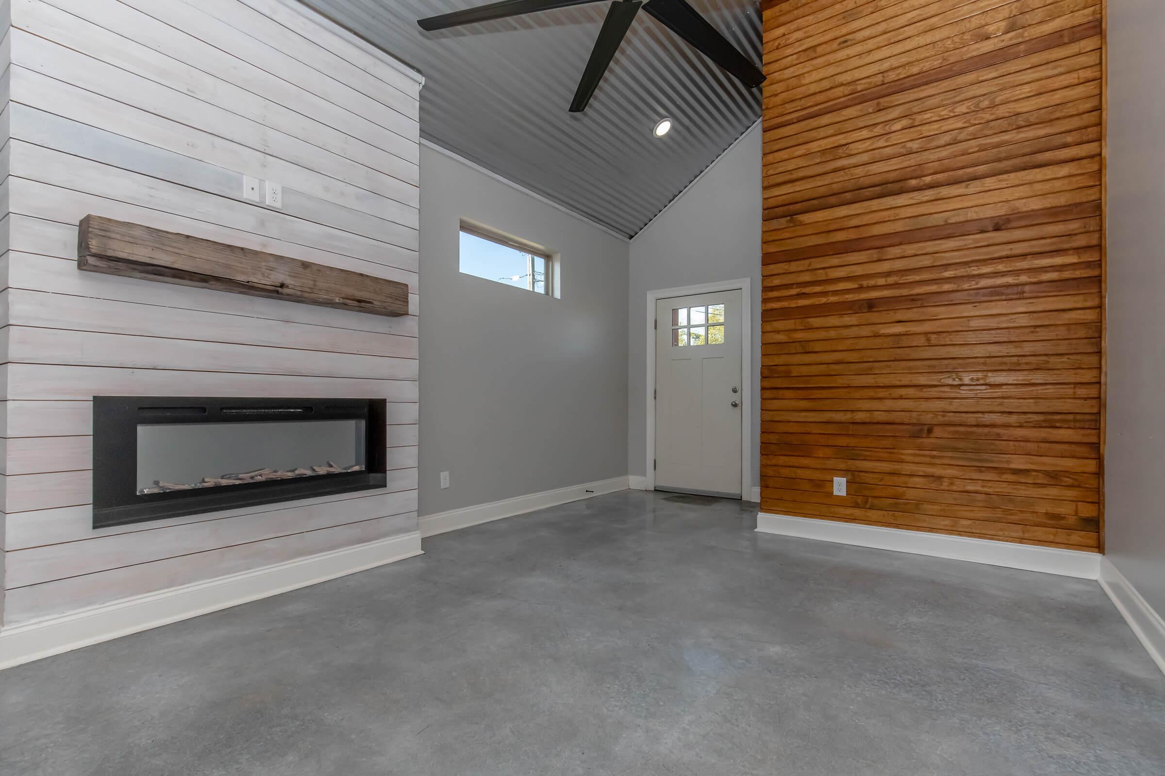 A modern interior space featuring a combination of white and wood-paneled walls. One side has a sleek electric fireplace built into a white wall, while the opposite wall is decorated with warm-toned wood paneling. The floor is polished concrete, and there’s a doorway leading outside. A ceiling fan is mounted above.