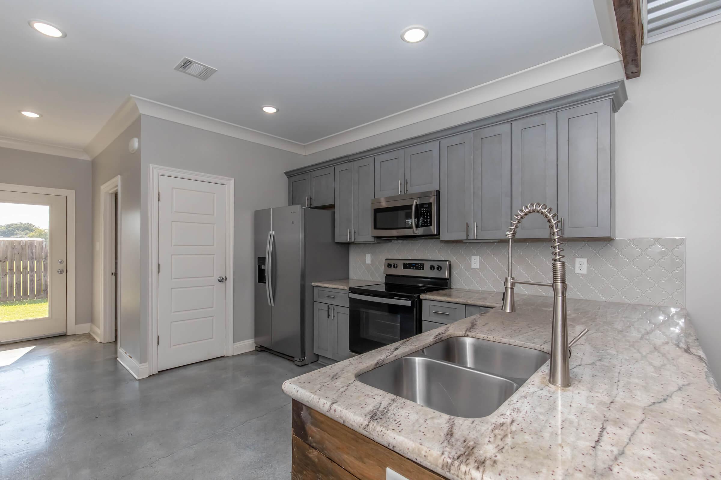 Modern kitchen featuring gray cabinetry, stainless steel appliances including a refrigerator and microwave, a granite countertop with a sink, and an open layout leading to a door with outdoor access. Natural light streams in through the windows, illuminating the minimalist design.