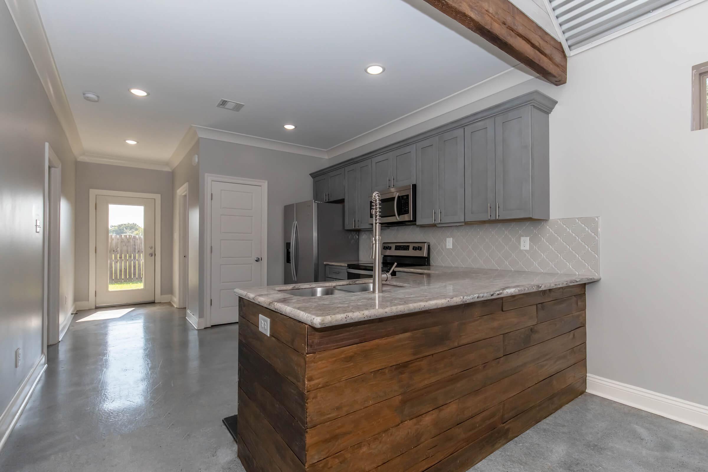 Modern kitchen featuring a central island with a wooden base and a granite countertop. Stainless steel appliances and gray cabinetry line the walls. The space has polished concrete floors and ample natural light coming from an entryway door leading outside.