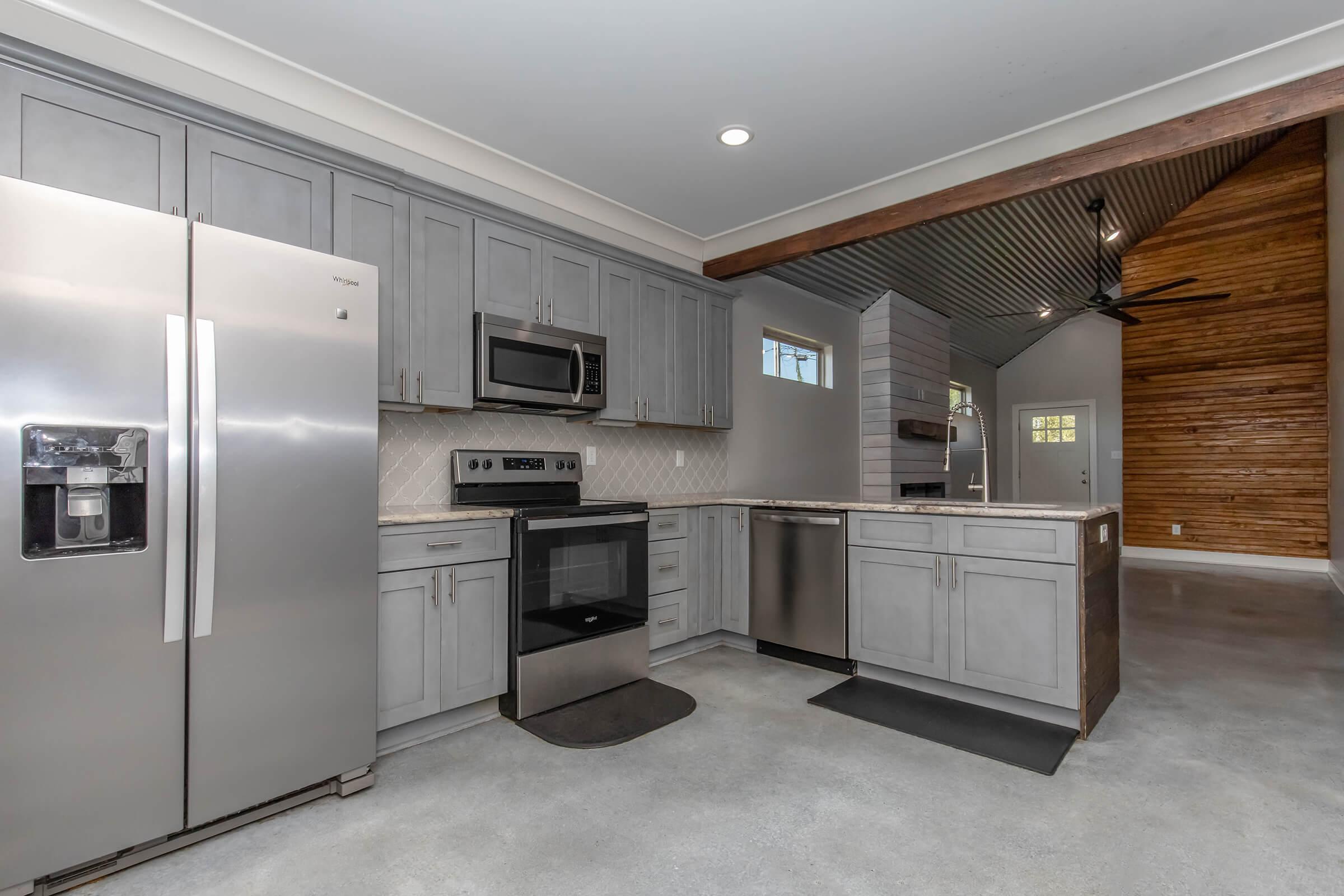 Modern kitchen featuring stainless steel appliances, including a refrigerator, microwave, and oven. Sleek gray cabinetry complements the light stone countertop, with a tiled backsplash. The open space has concrete floors and wooden accents, showcasing a contemporary design.