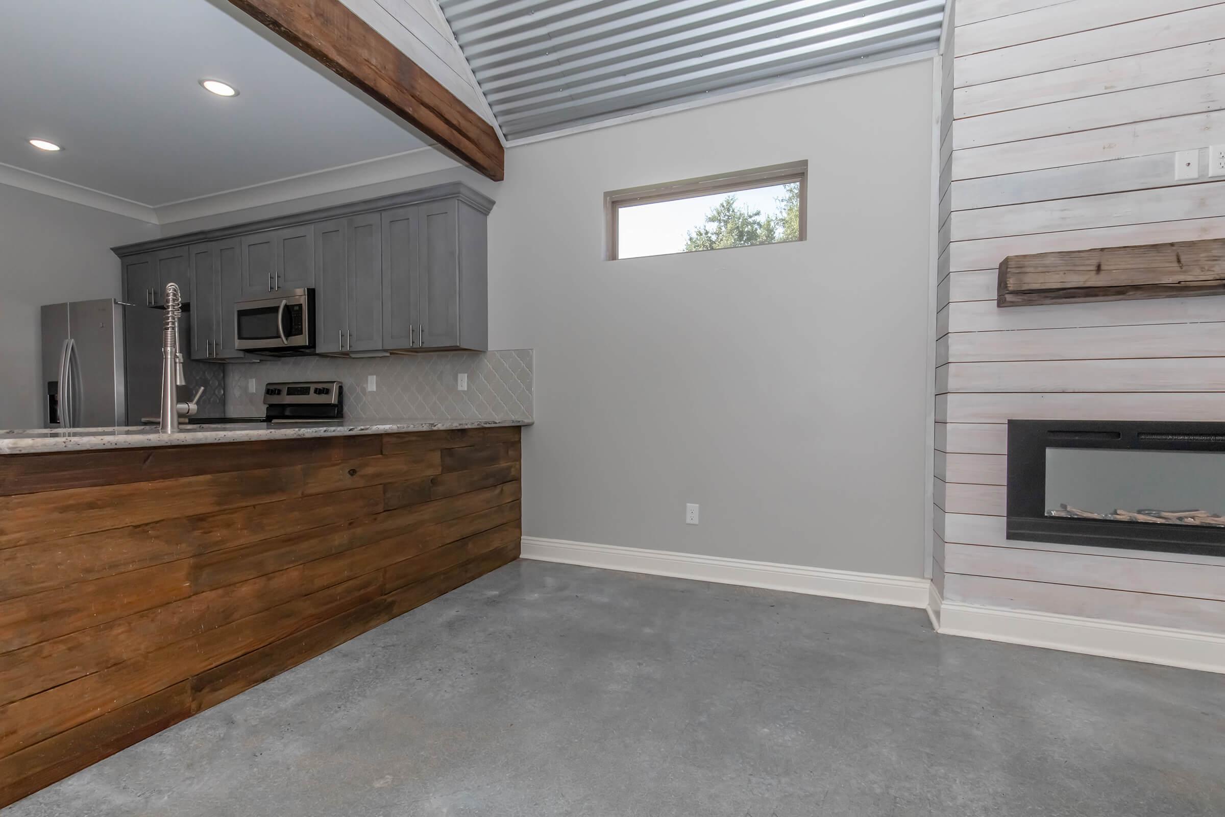 Interior of a modern living space featuring a kitchen with gray cabinets, stainless steel appliances, and a granite countertop. The adjacent wall has a rustic wooden element and a minimalist fireplace, while large windows provide natural light and a view of greenery outside.