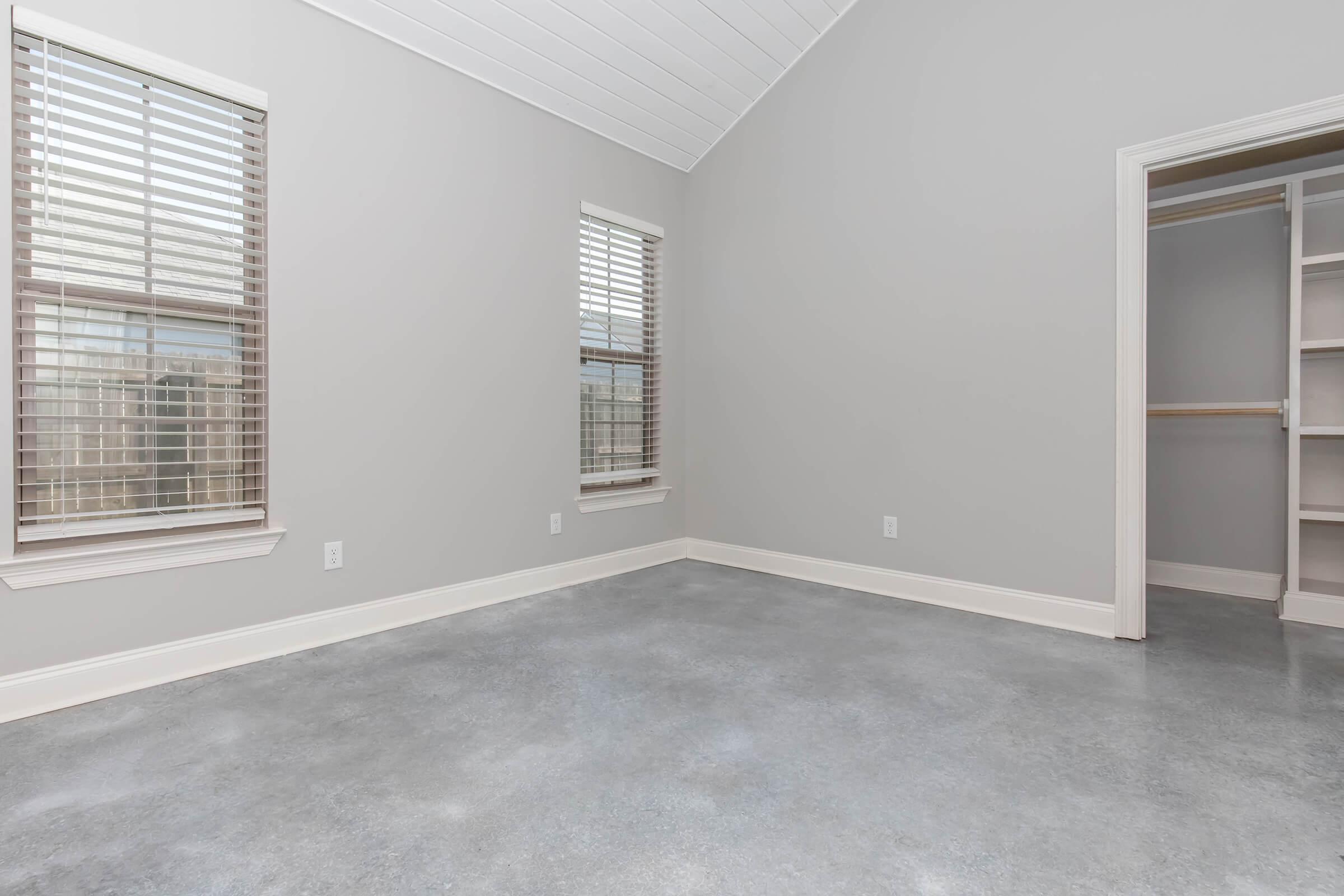 A spacious, empty room with gray walls and a high ceiling. There are two windows with blinds allowing natural light. The floor is polished concrete, and there’s a built-in closet on the right with shelves visible. The overall atmosphere is modern and minimalistic.