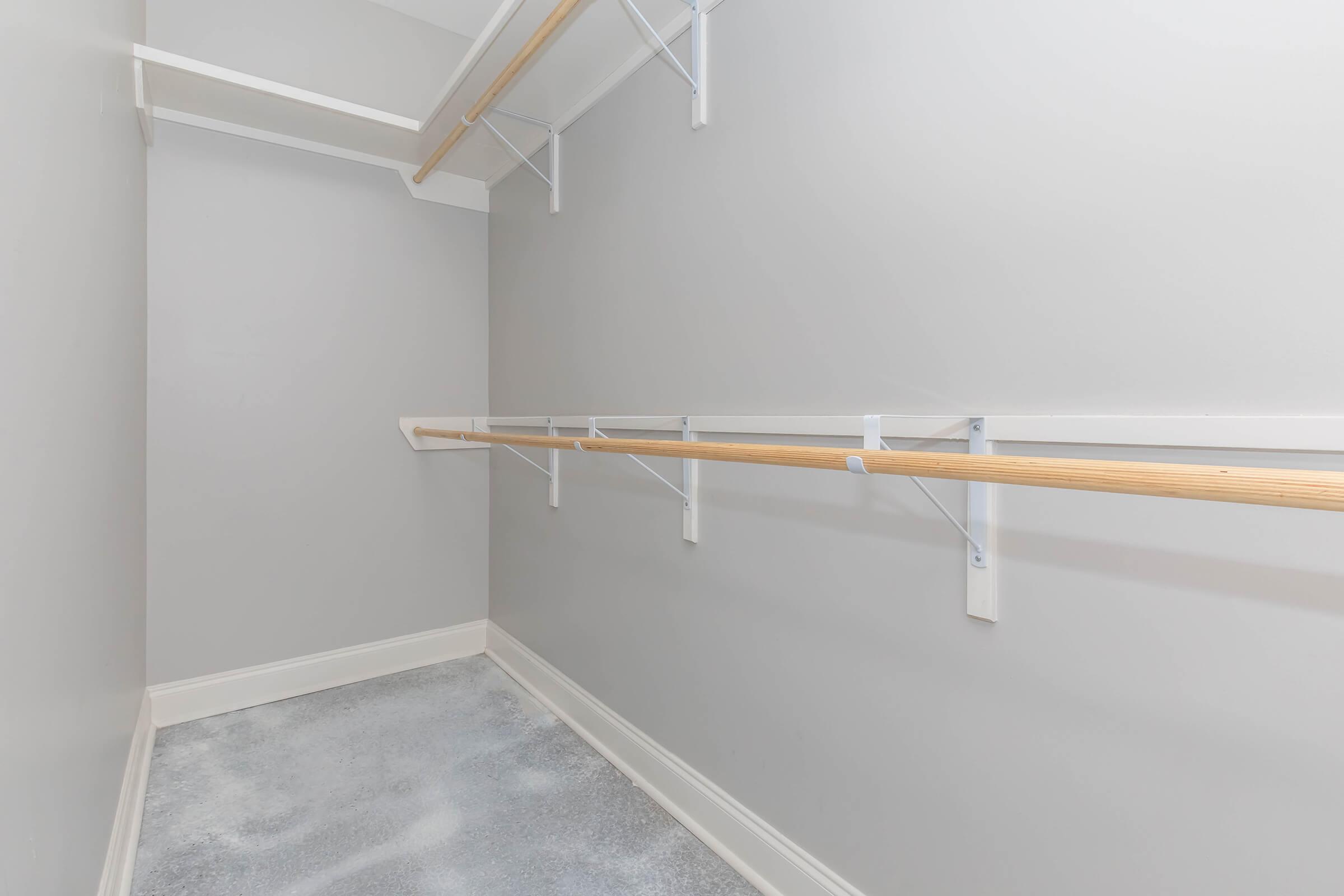 Empty walk-in closet with gray walls and a concrete floor, featuring two horizontal wooden rods for hanging clothes. The space is well-lit and unobstructed, showcasing a minimalist design without any furnishings or items.