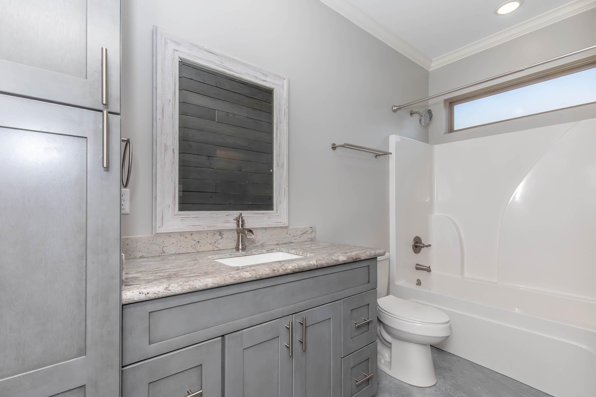 A modern bathroom featuring a gray vanity with a granite countertop, a white sink, and an elegant mirror. The space includes a white bathtub and shower combination, gray walls, and sleek fixtures, with natural light coming through a small window above the tub.