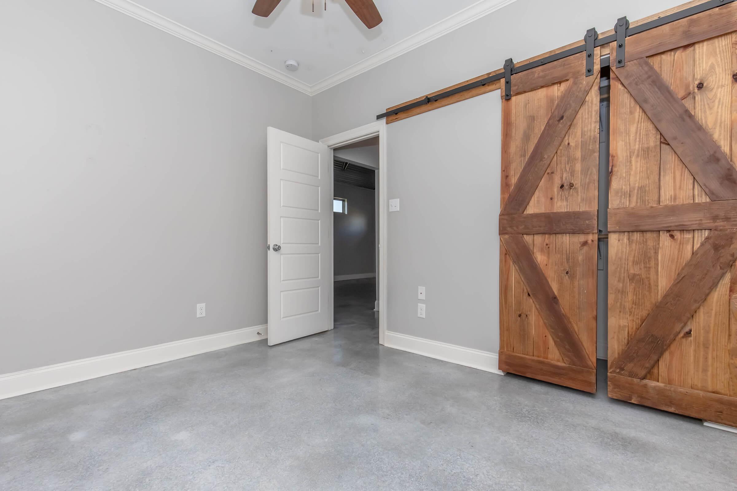 Interior view of a modern room featuring a light gray wall, polished concrete flooring, and a wooden sliding barn door. A white door is slightly open, leading to another space. The room has a ceiling fan and a clean, minimalist aesthetic, creating a bright and inviting atmosphere.