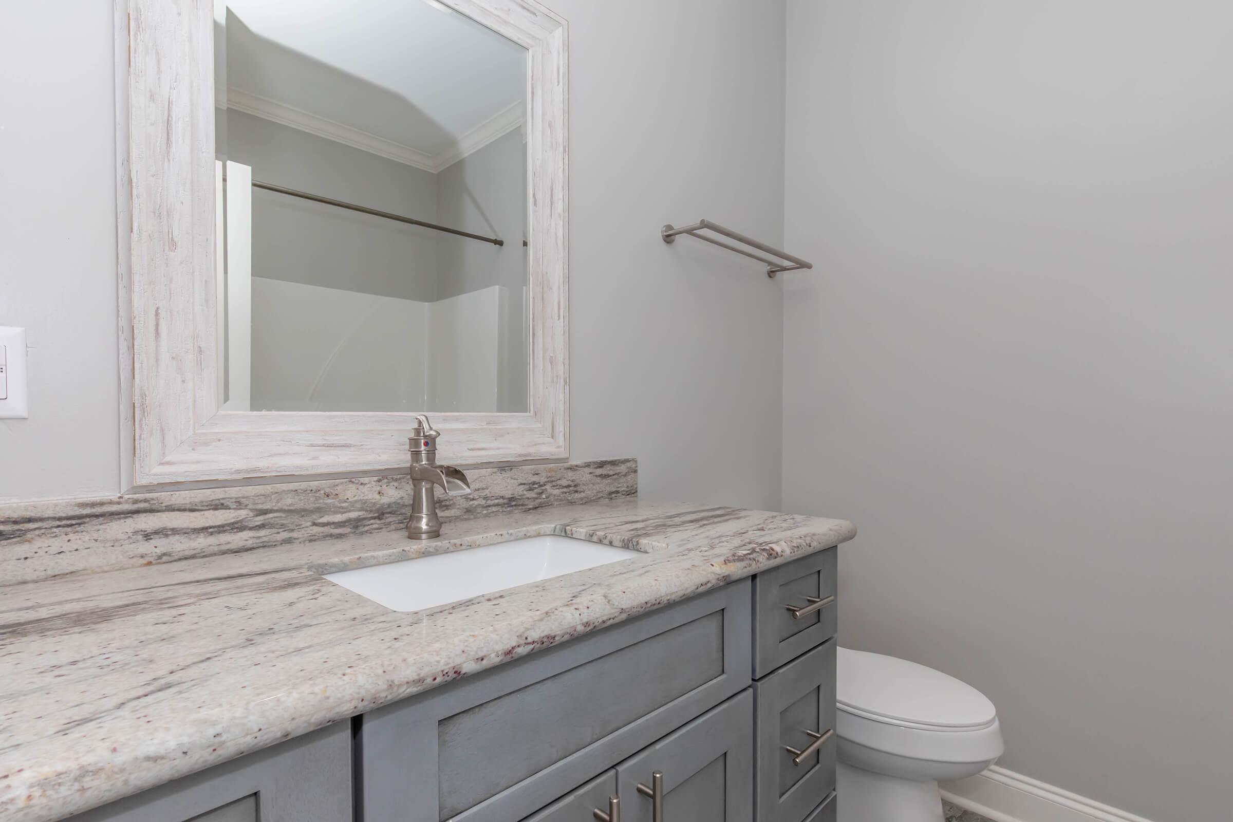 A modern bathroom with light gray walls. The vanity features a granite countertop with a sink, silver faucet, and drawers. A framed mirror hangs above the sink, and a towel bar is mounted on the wall. A toilet is visible to the right, completing the clean, minimalistic design.