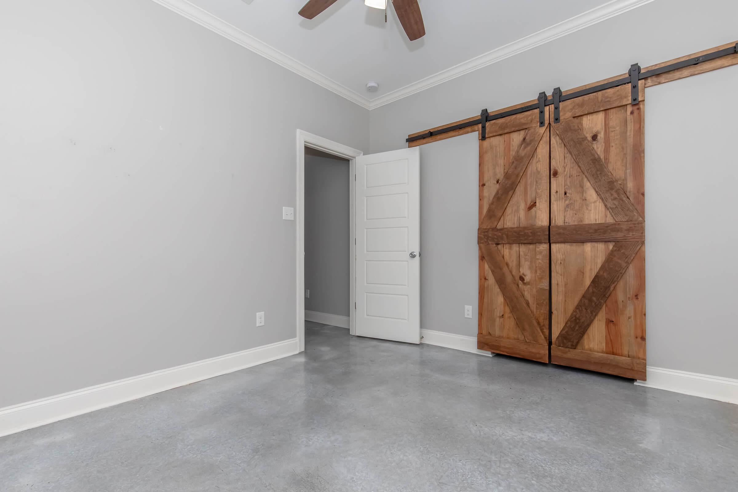 Empty room with a gray color scheme, featuring a concrete floor and white ceiling. A wooden sliding barn door with a diagonal pattern is mounted on the wall, alongside a standard closed door leading to another room. Bright lighting illuminates the space.
