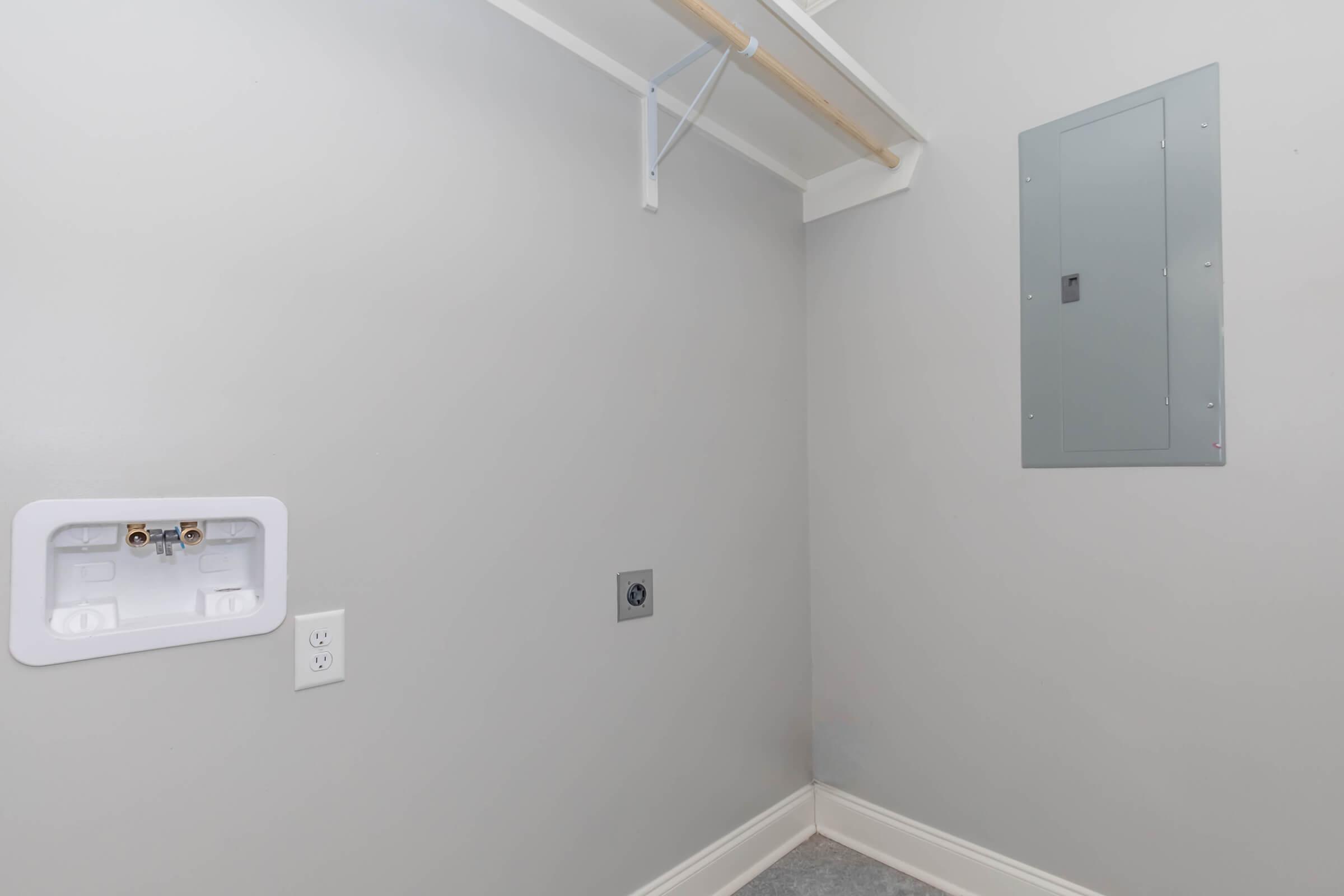 A well-lit laundry room with light gray walls, featuring a mounted electrical panel, a white utility outlet, and plumbing connections for a washing machine. Shelving is visible above, with a clean, uncluttered floor and a neutral color scheme, creating a modern and functional space.