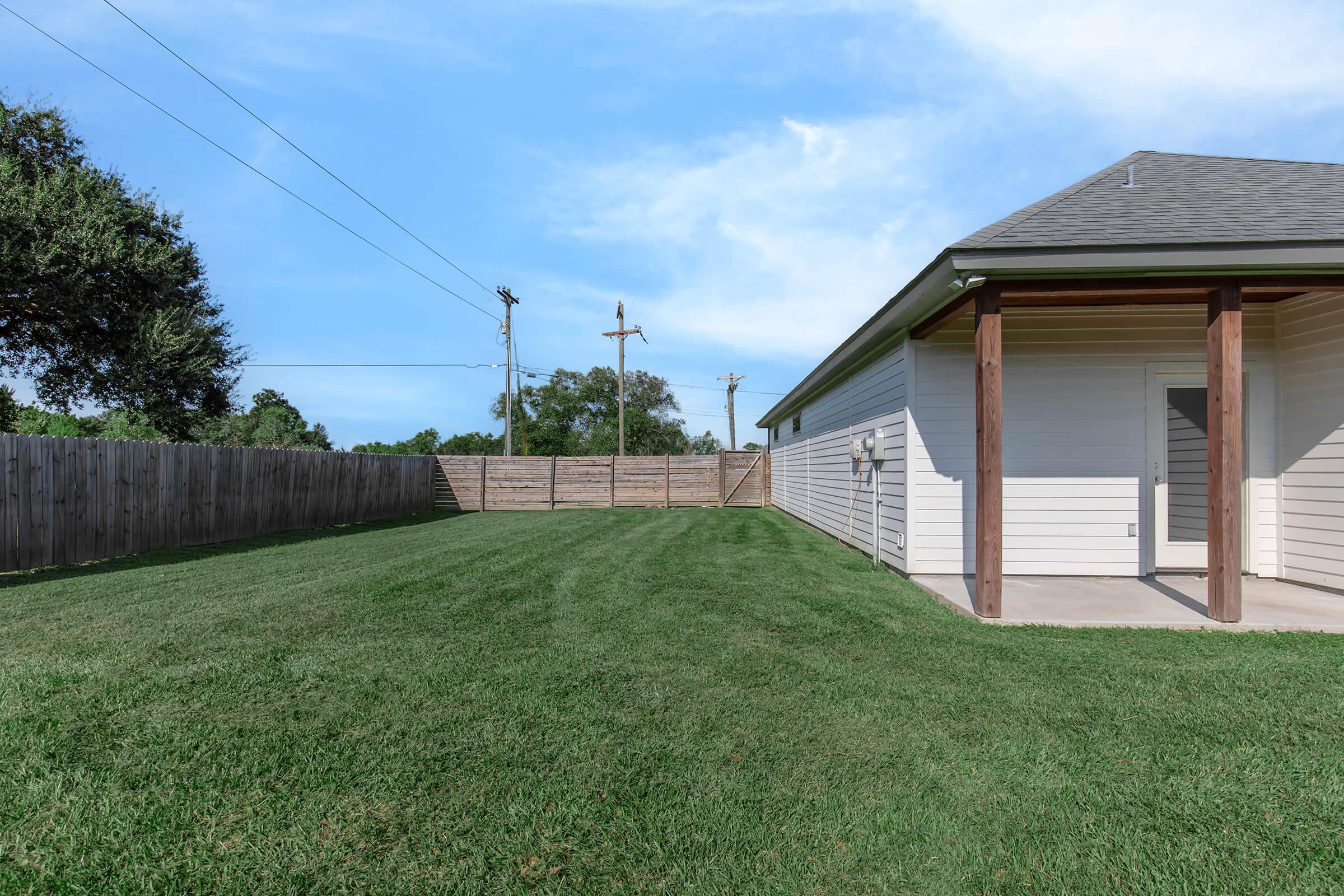 A well-maintained backyard with lush green grass, featuring a clear blue sky overhead and a simple gray house on the right. The yard is enclosed by a wooden fence, with power lines visible in the background. A patio area is shown next to the house, providing an inviting outdoor space.
