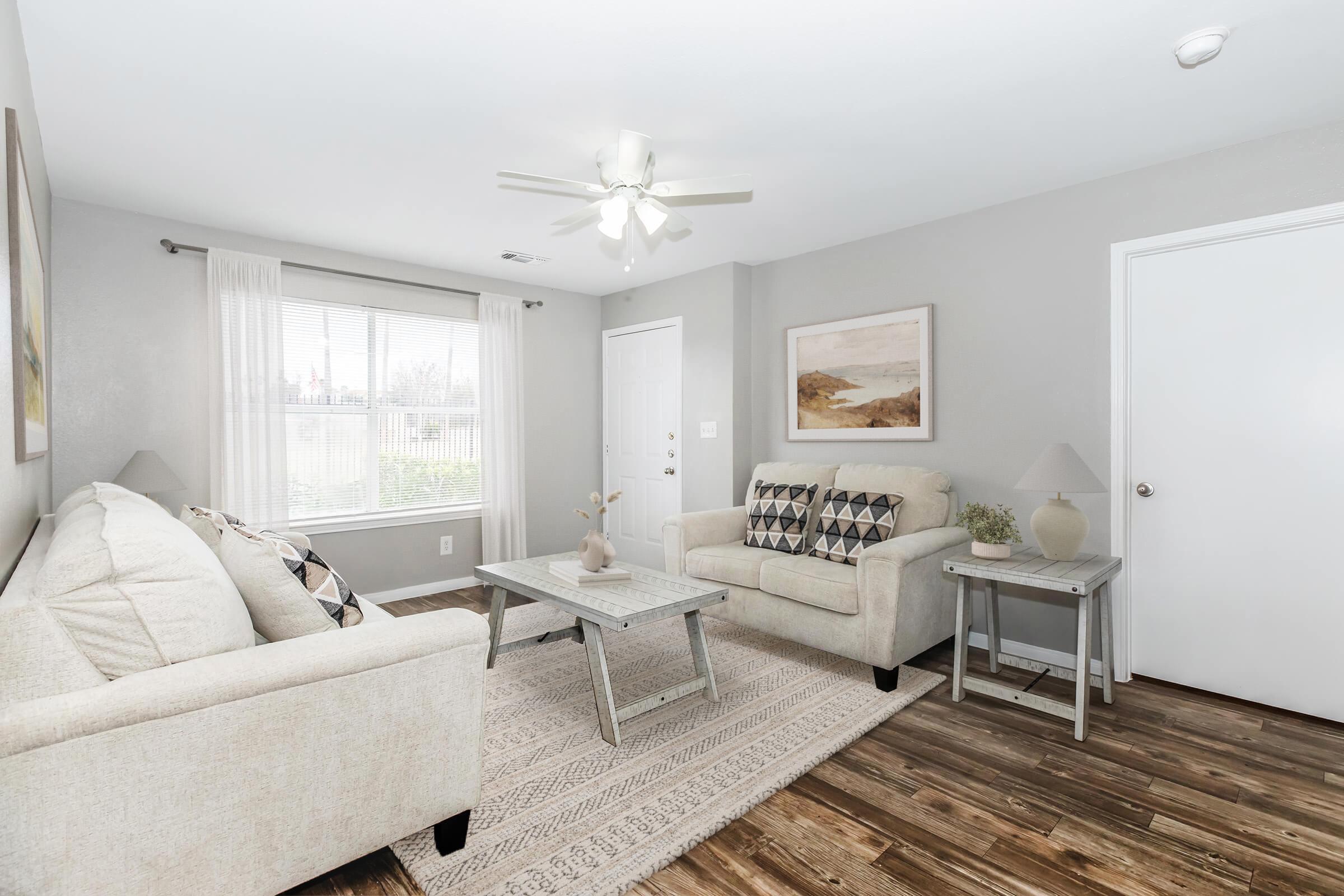 Cozy living room featuring a light-colored sofa and armchair, a wooden coffee table, and a textured area rug. Natural light streams in through a window with white curtains, illuminating the space. A wall art piece and a small lamp add decorative touches to the inviting atmosphere.