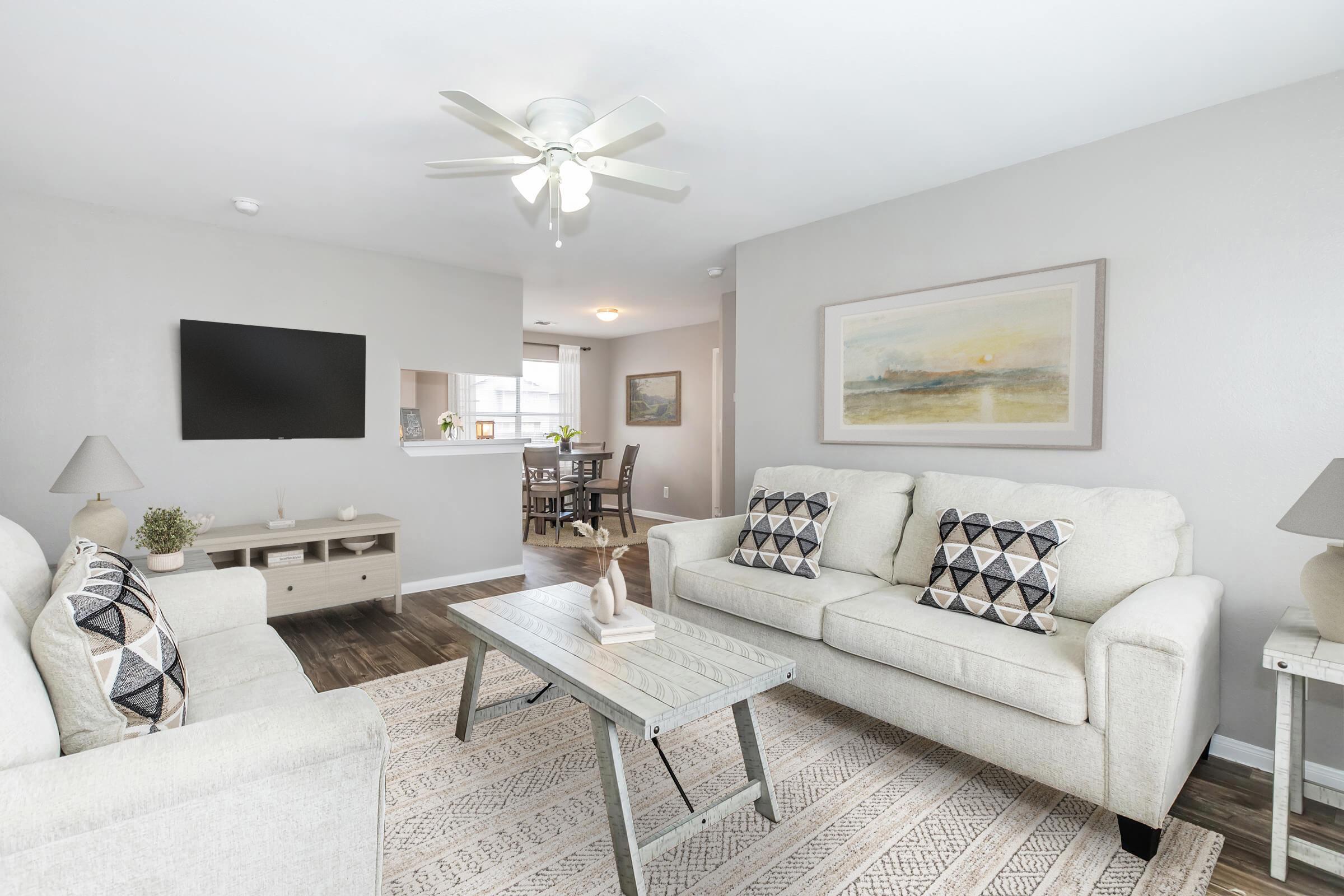A bright, modern living room featuring two light-colored sofas with patterned cushions, a gray coffee table, and a decorative piece on top. There’s a flat-screen TV mounted on the wall and an open kitchen area in the background. The space is illuminated by natural light, creating a welcoming atmosphere.