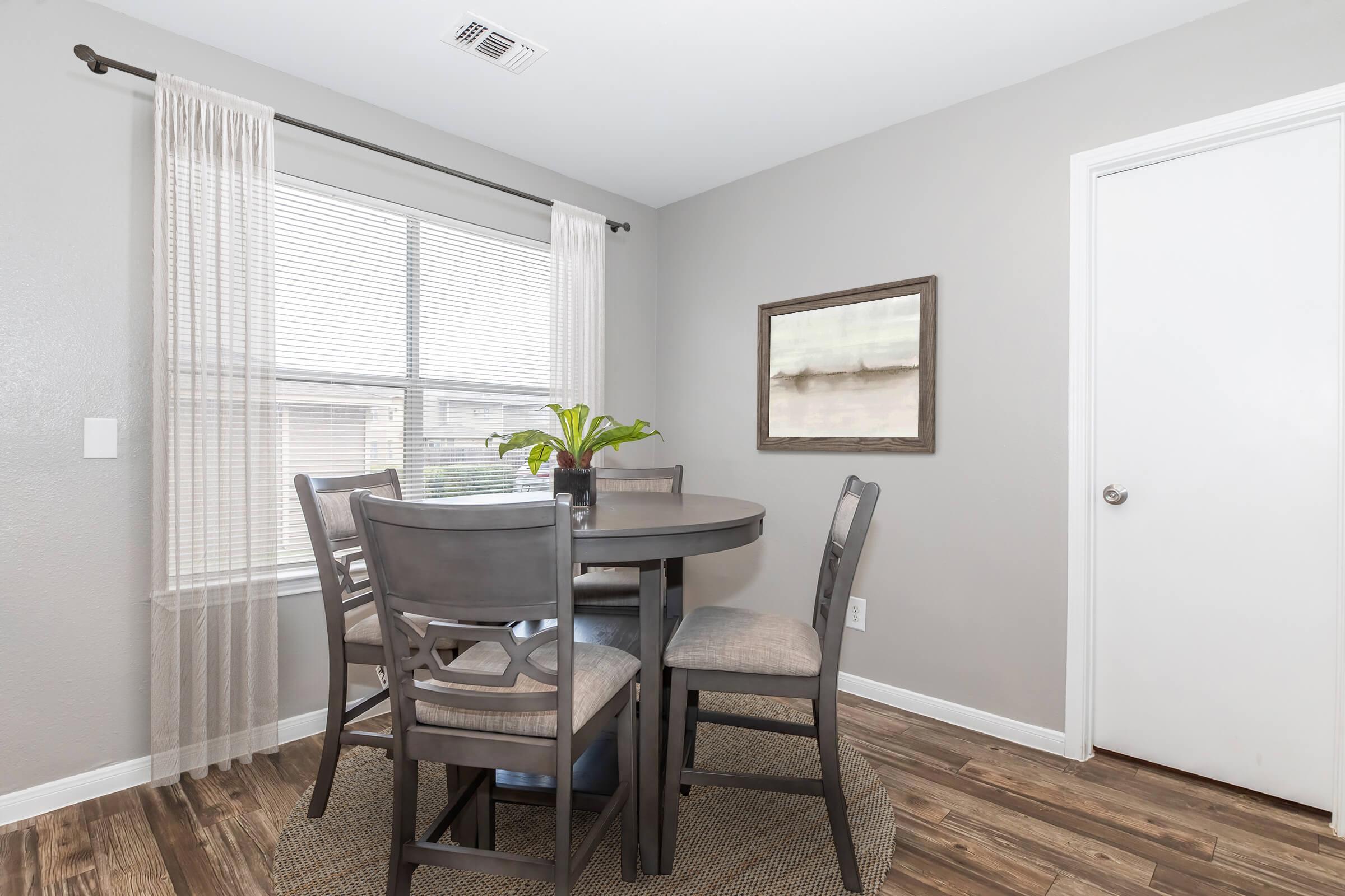 A dining area featuring a small round table surrounded by four gray chairs. A stylish plant sits at the center of the table. The room includes a window with sheer curtains, a framed artwork on the wall, and a door leading to another space. The floor is covered with wooden laminate.