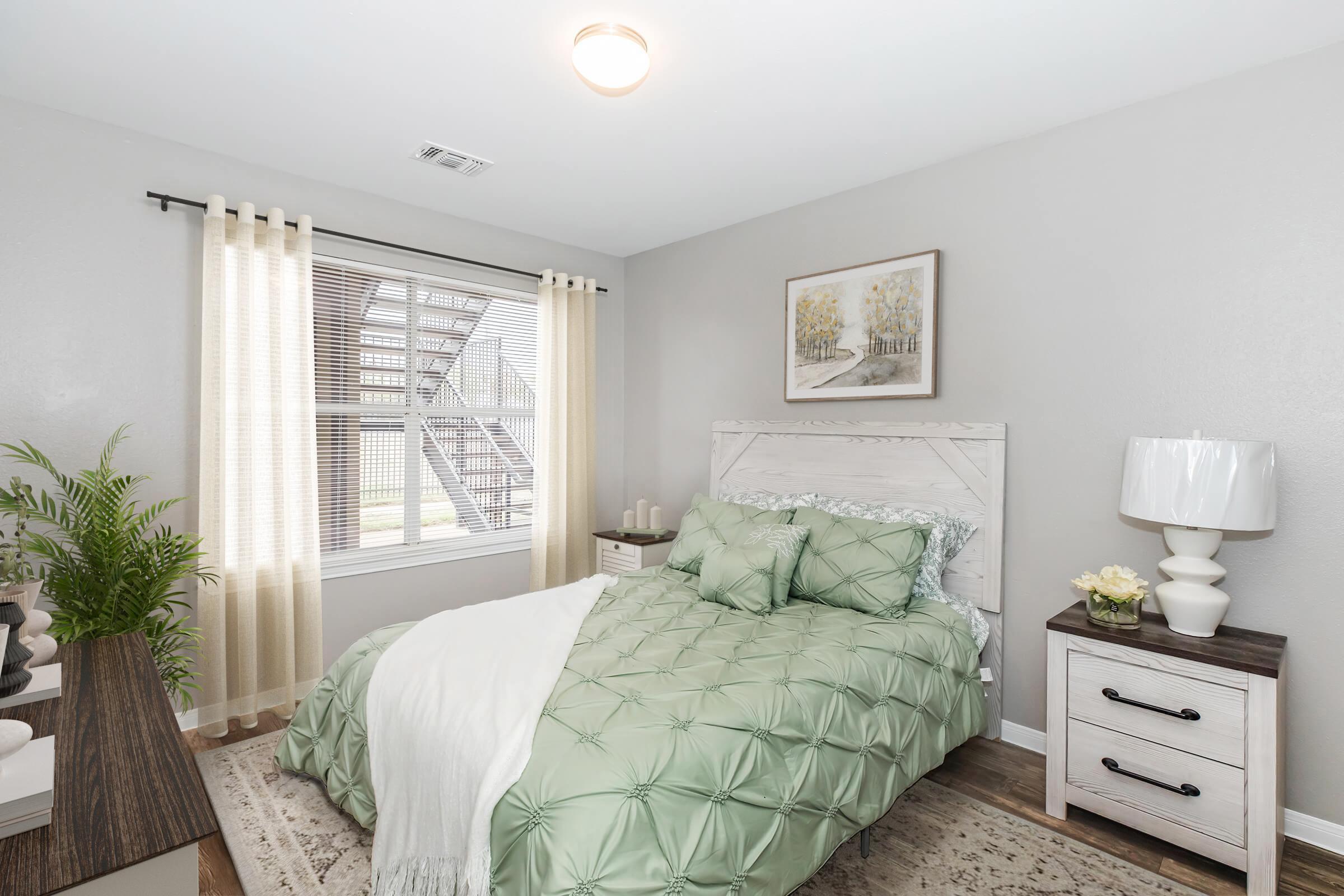 Cozy bedroom featuring a light green quilted bedspread, a nightstand with a lamp, and decorative artwork on the wall. Natural light enters through a window adorned with sheer curtains, complemented by a potted plant and a decorative rug on the floor.