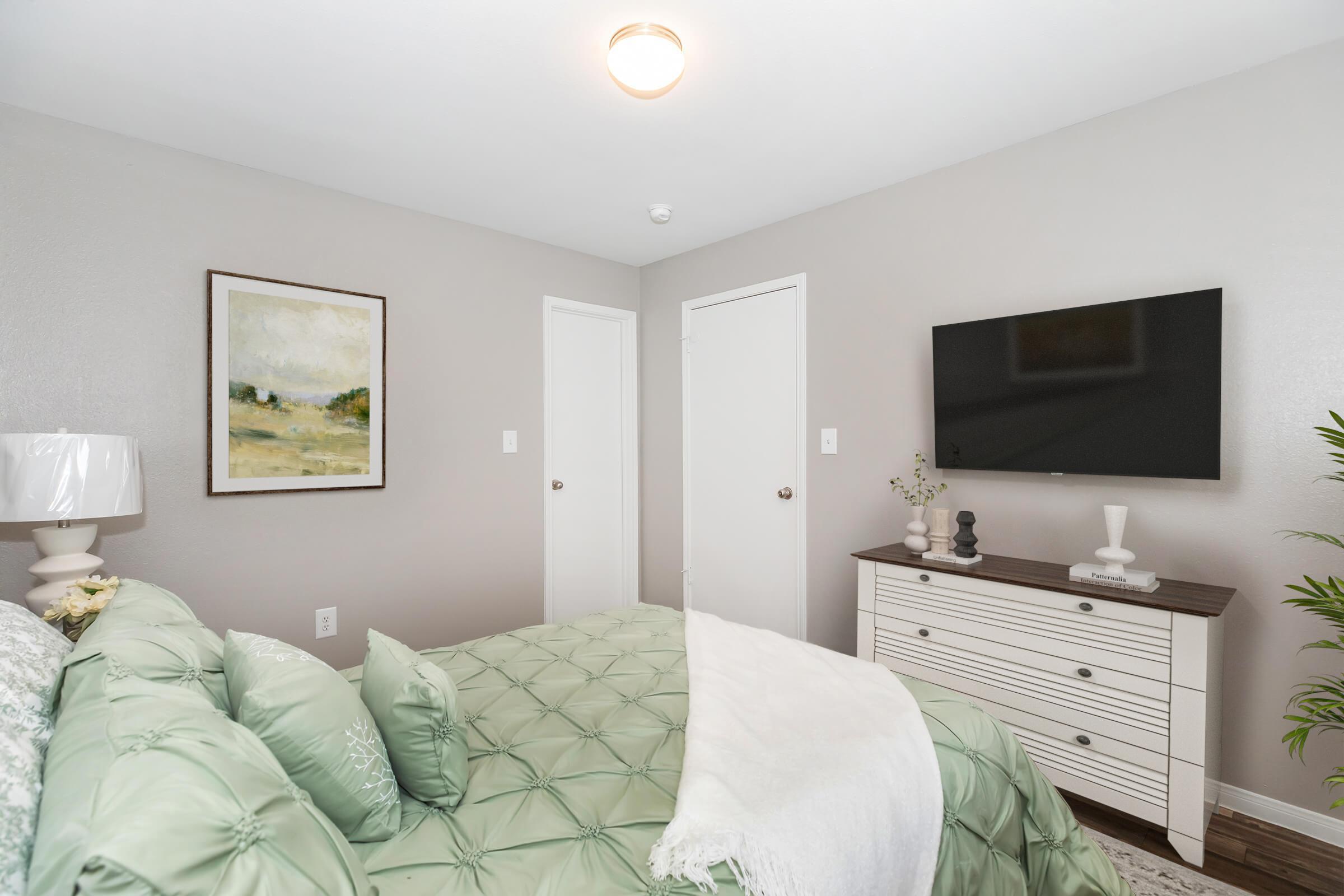 A cozy bedroom featuring a neatly made bed with green bedding, a lamp on the nightstand, an abstract painting on the wall, a television mounted above a white dresser, and two closed doors. The walls are painted a soft gray, creating a calming atmosphere. A plant adds a touch of greenery.