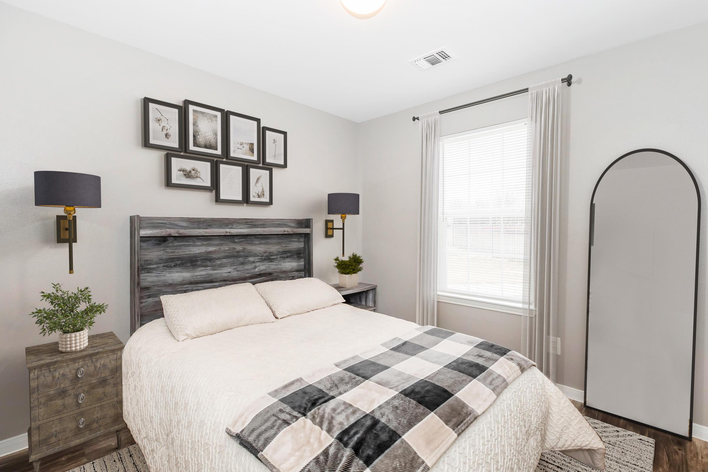 A cozy bedroom featuring a wooden bed frame with light bedding and a plaid throw. There are two wall-mounted lamps beside the bed, a set of framed artworks above, and a small bedside table with a plant. A full-length mirror is positioned near the window, which has light curtains letting in natural light.