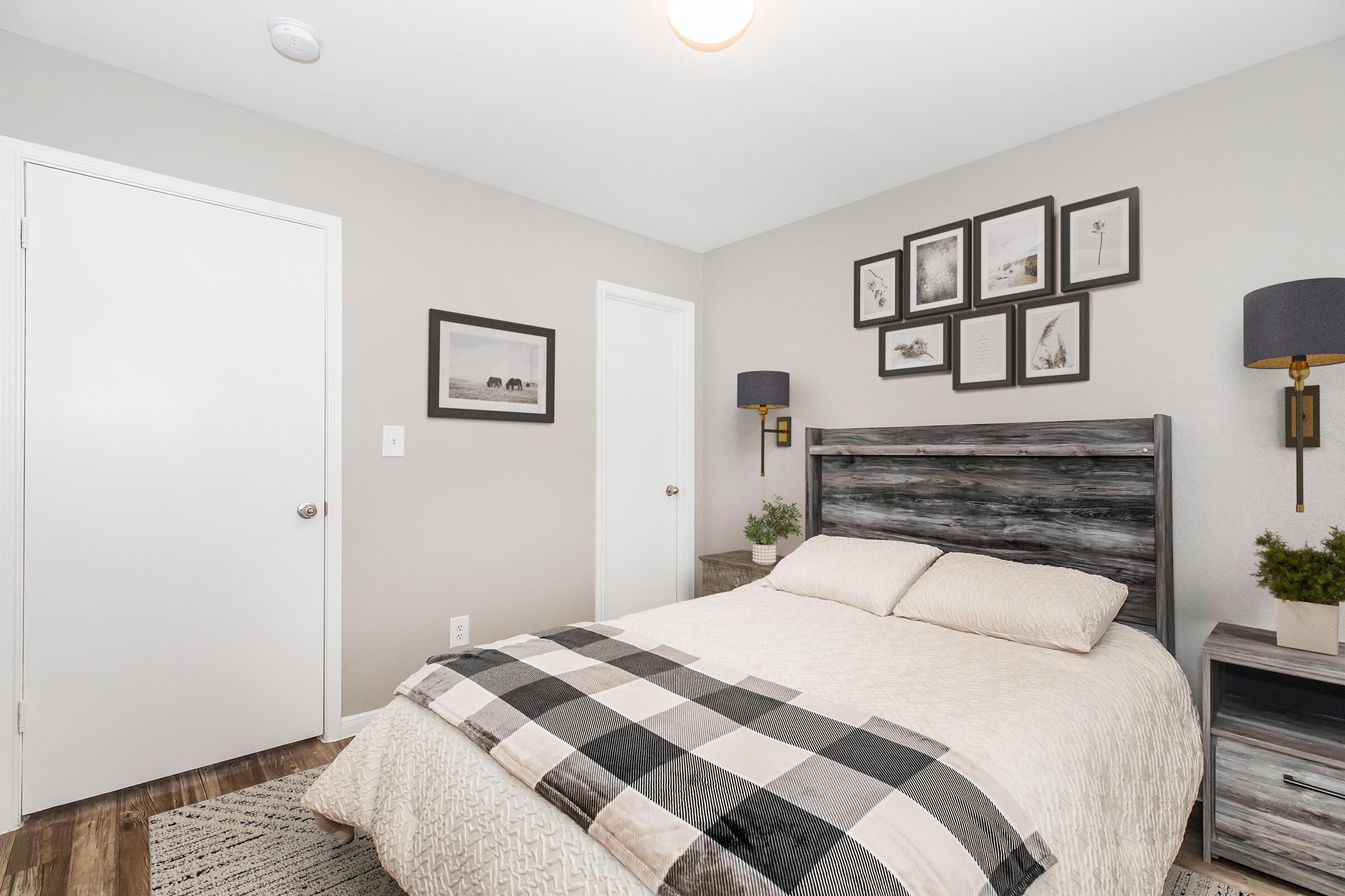A cozy bedroom featuring a wooden bed with a beige and black checkered throw, accented by gray walls. Wall art consists of framed pictures, while bedside lamps add a soft ambiance. A closed white door and a white closet door are visible, along with small potted plants on the nightstands, enhancing the inviting atmosphere.