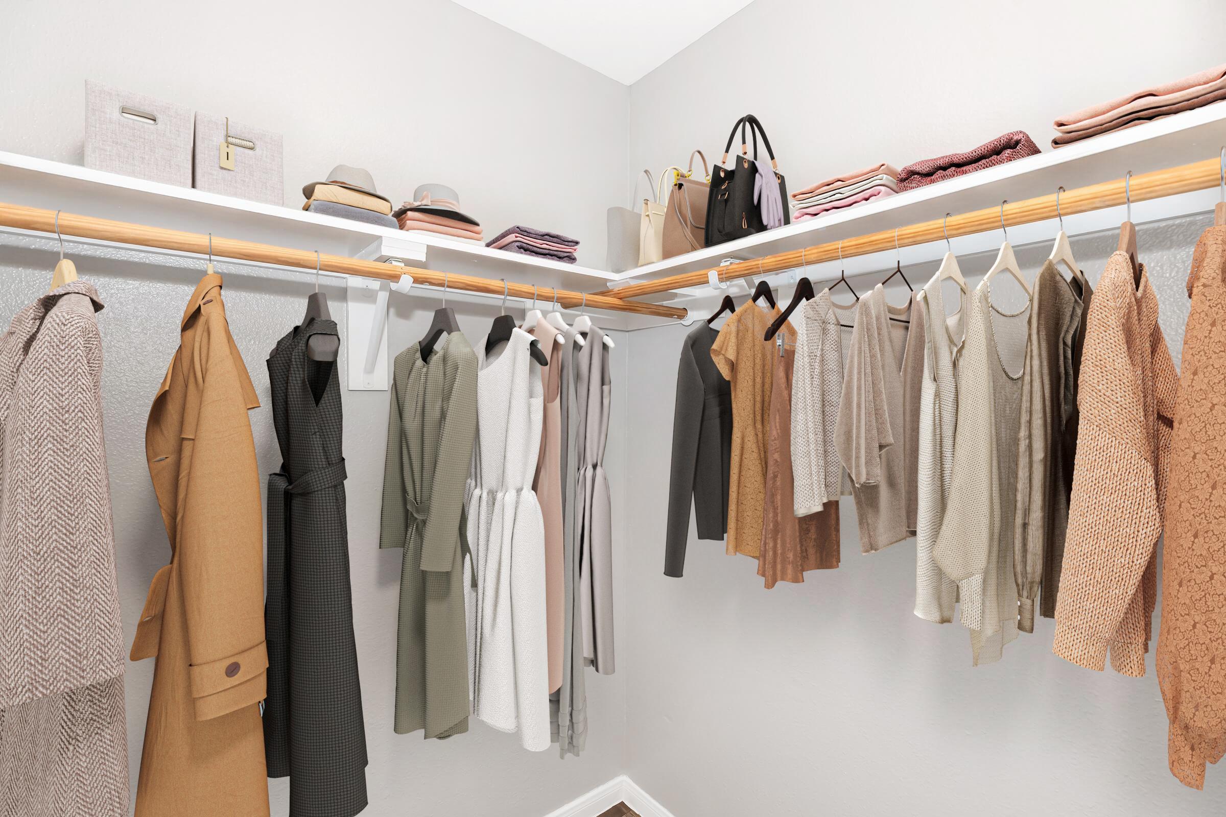 A neatly organized walk-in closet featuring various clothing items on hangers, including dresses, coats, and sweaters in neutral tones. There are also neatly folded items on the shelves above, along with several handbags and a hat, all set against a light gray wall.